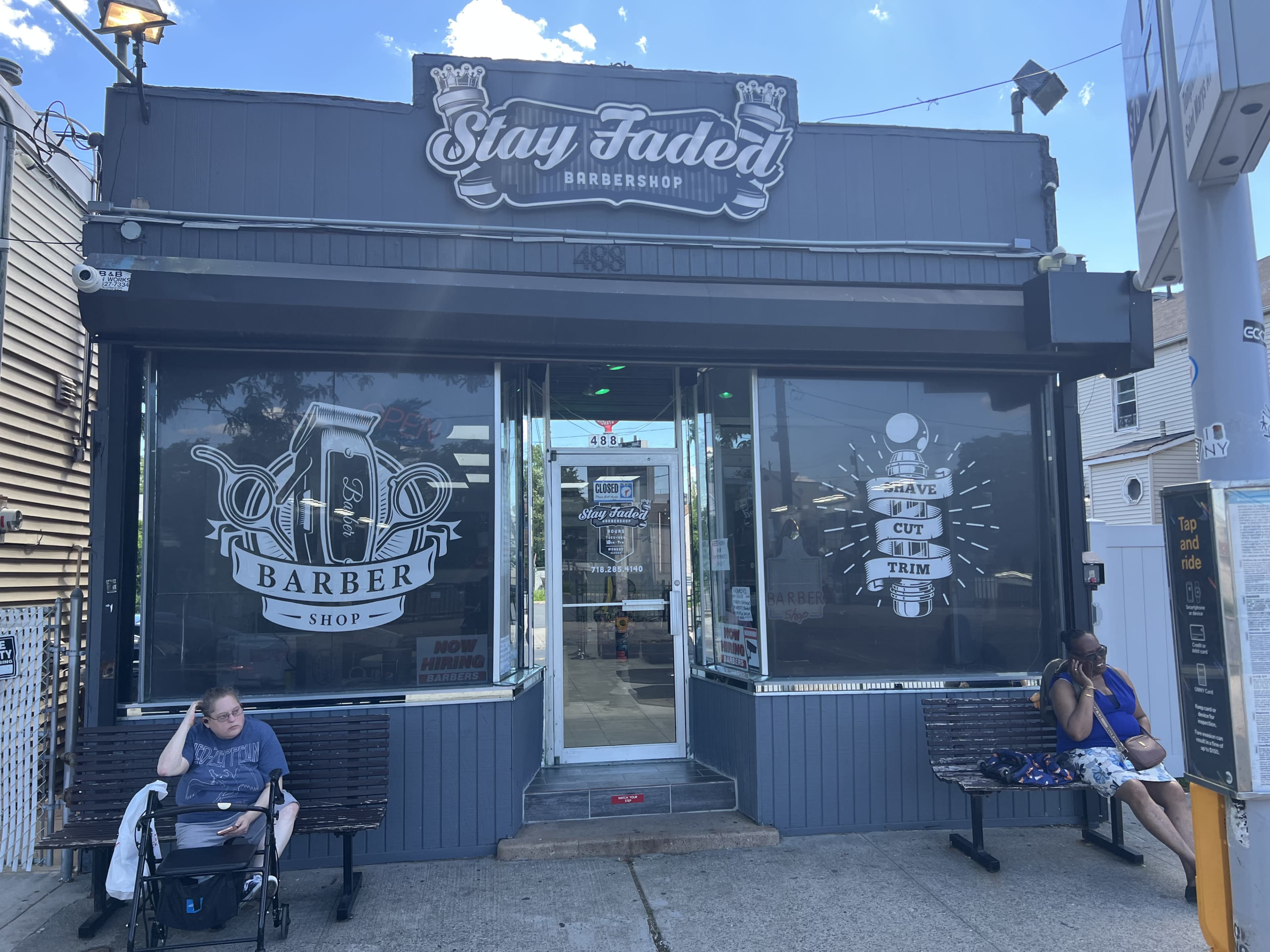 The image shows the storefront of a barbershop named "Stay Faded," with large graphic windows displaying barber-related artwork and two people sitting on benches outside.