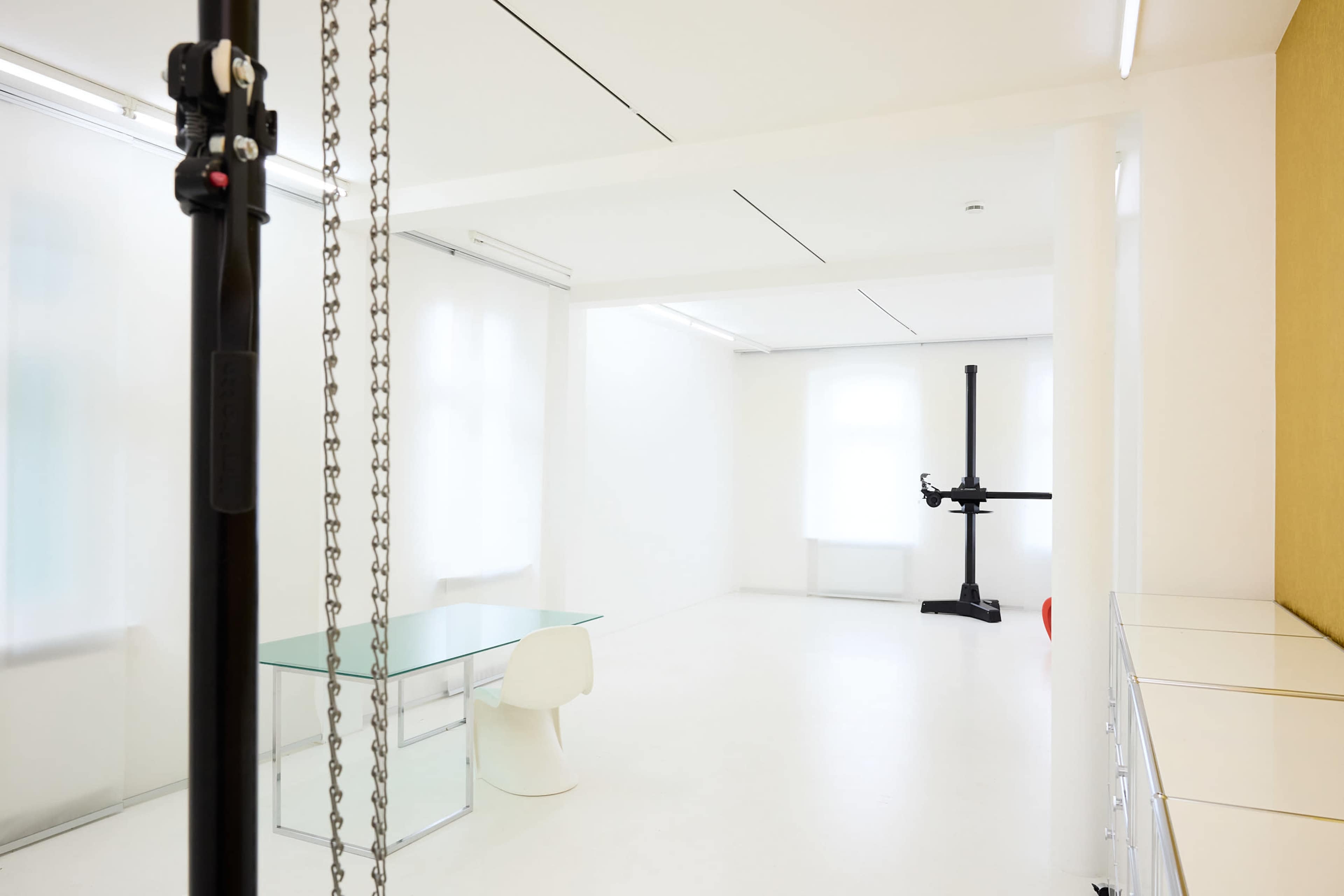 The image shows a minimalist white room featuring a glass desk and a simple white chair, with a mechanical structure in the corner and large windows allowing natural light.