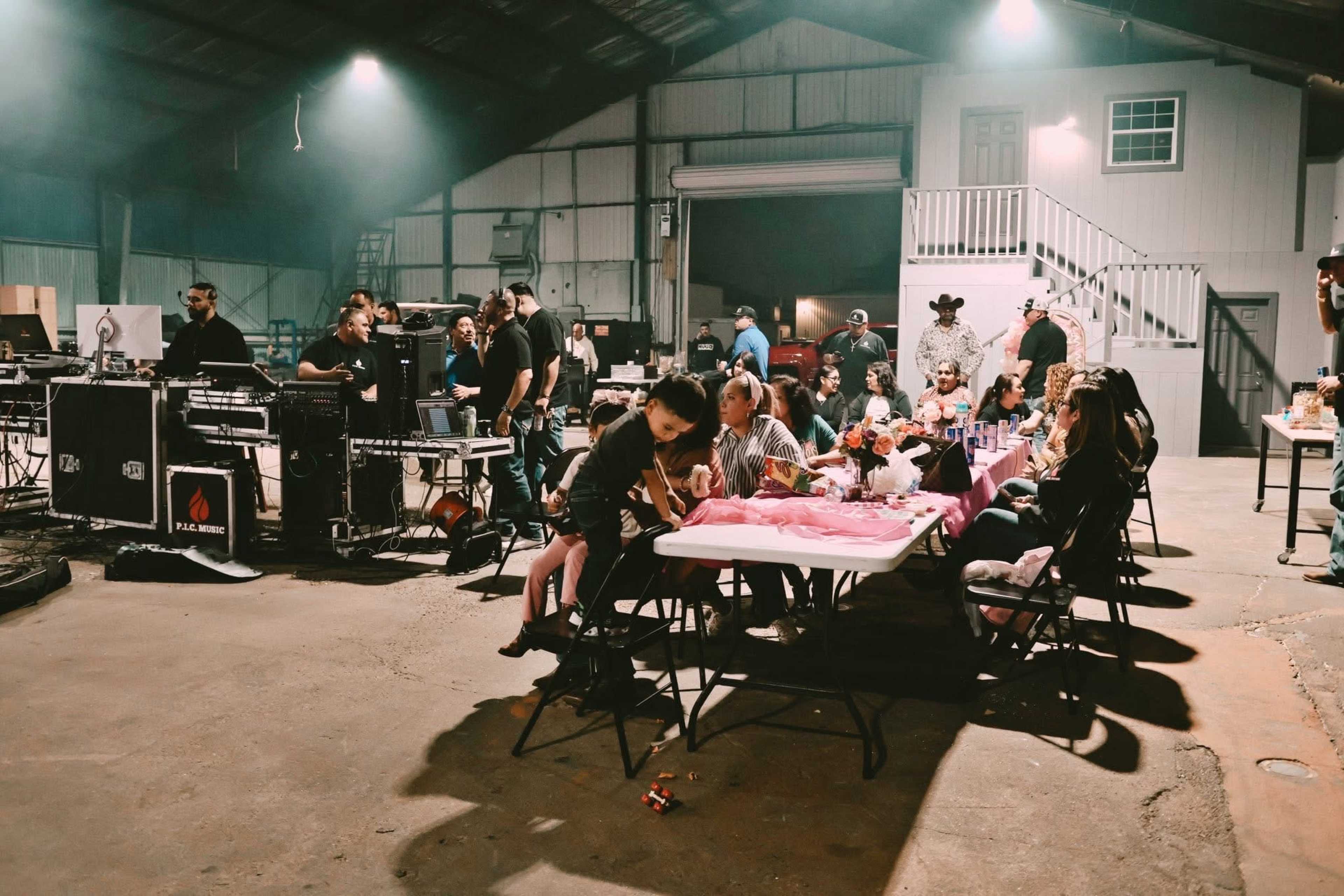 A group of people gathers around tables in a large indoor space, with equipment and staging visible in the background.