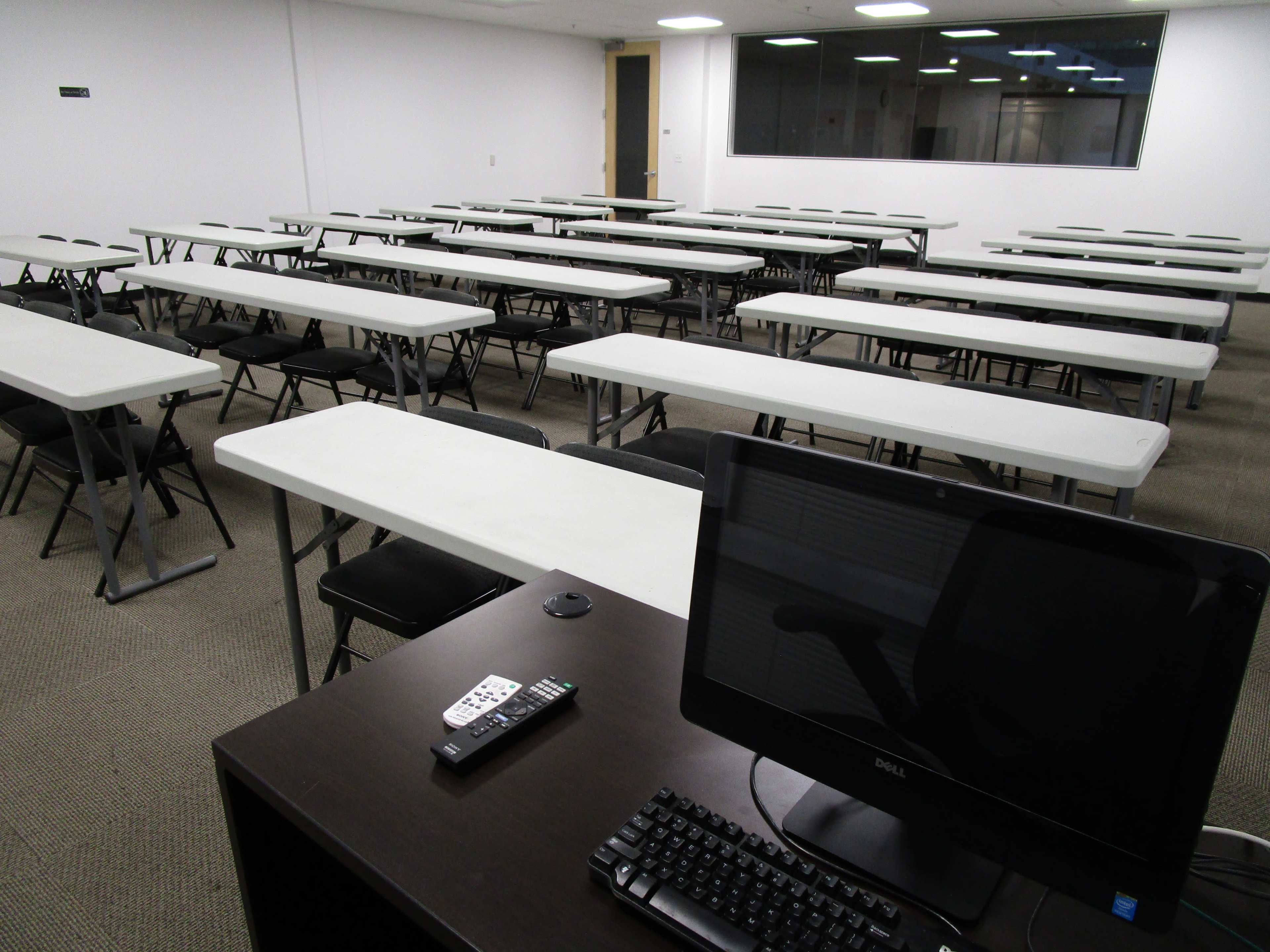 A classroom or meeting room with multiple rows of folding tables and chairs, alongside a desk featuring a computer and remote control.