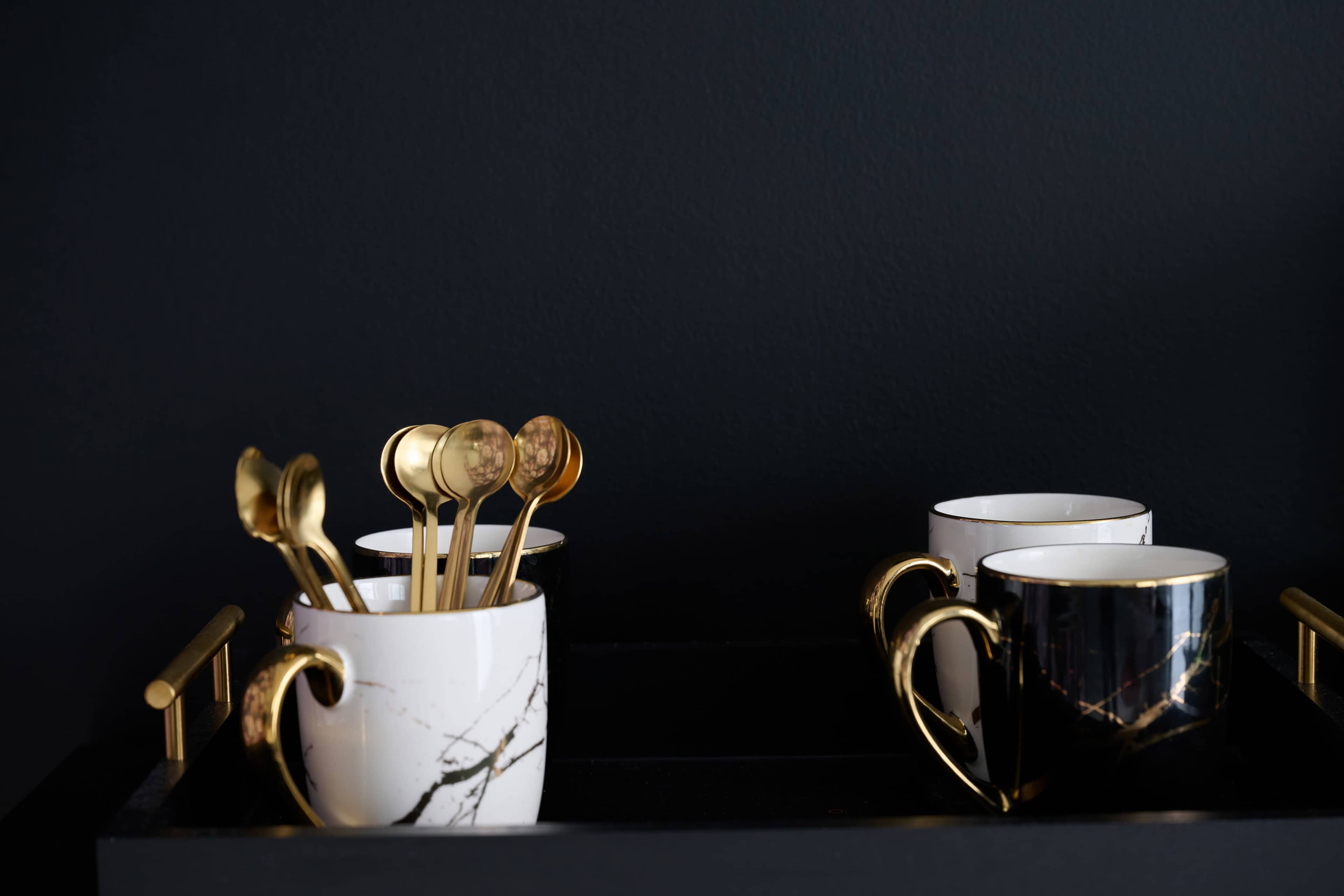 A black tray holds two mugs, one white with a marble pattern and one black, alongside gold spoons in a decorative white cup.