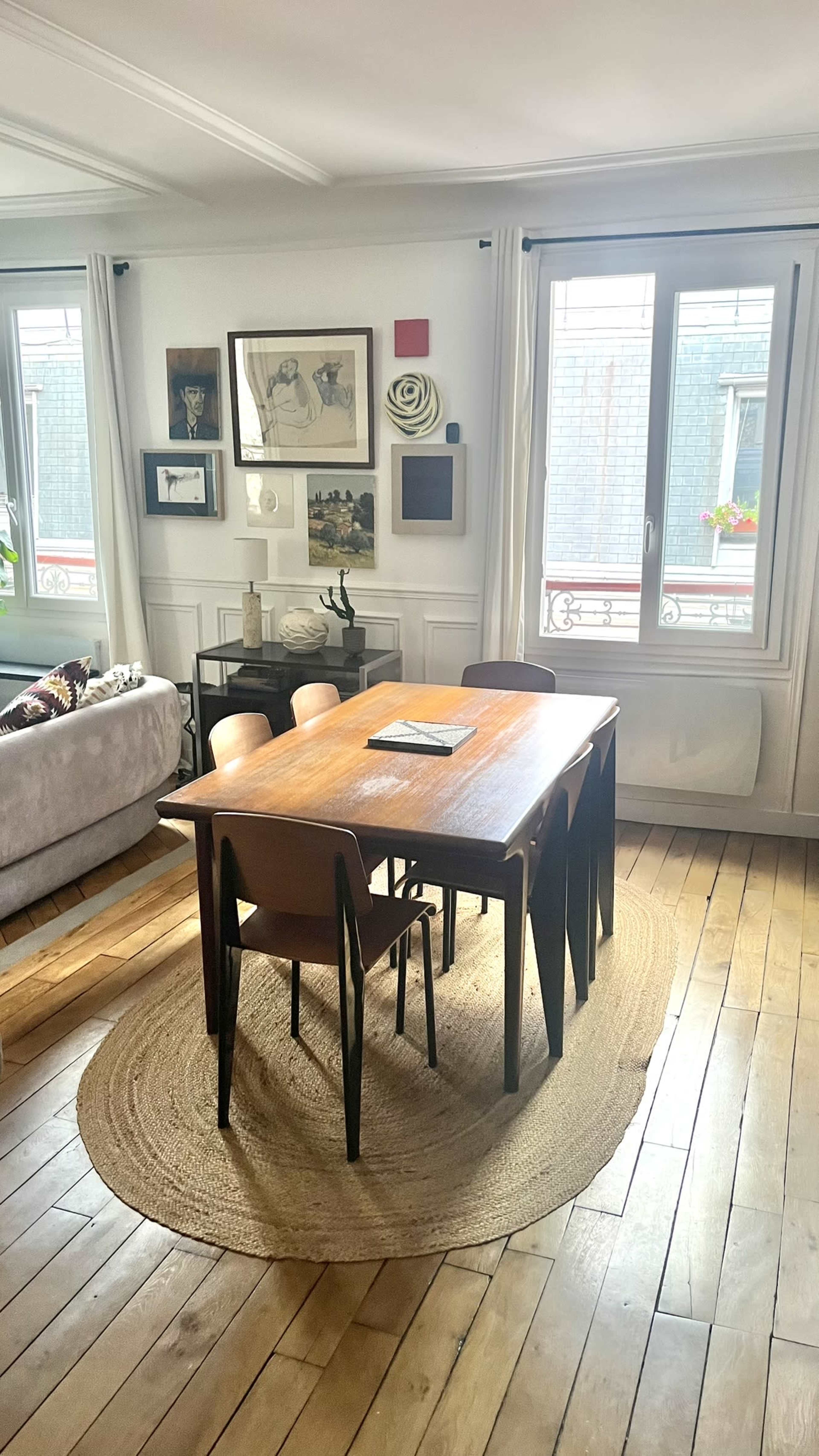 A wooden dining table surrounded by chairs is situated on a round rug in a well-lit room featuring framed artwork and windows.