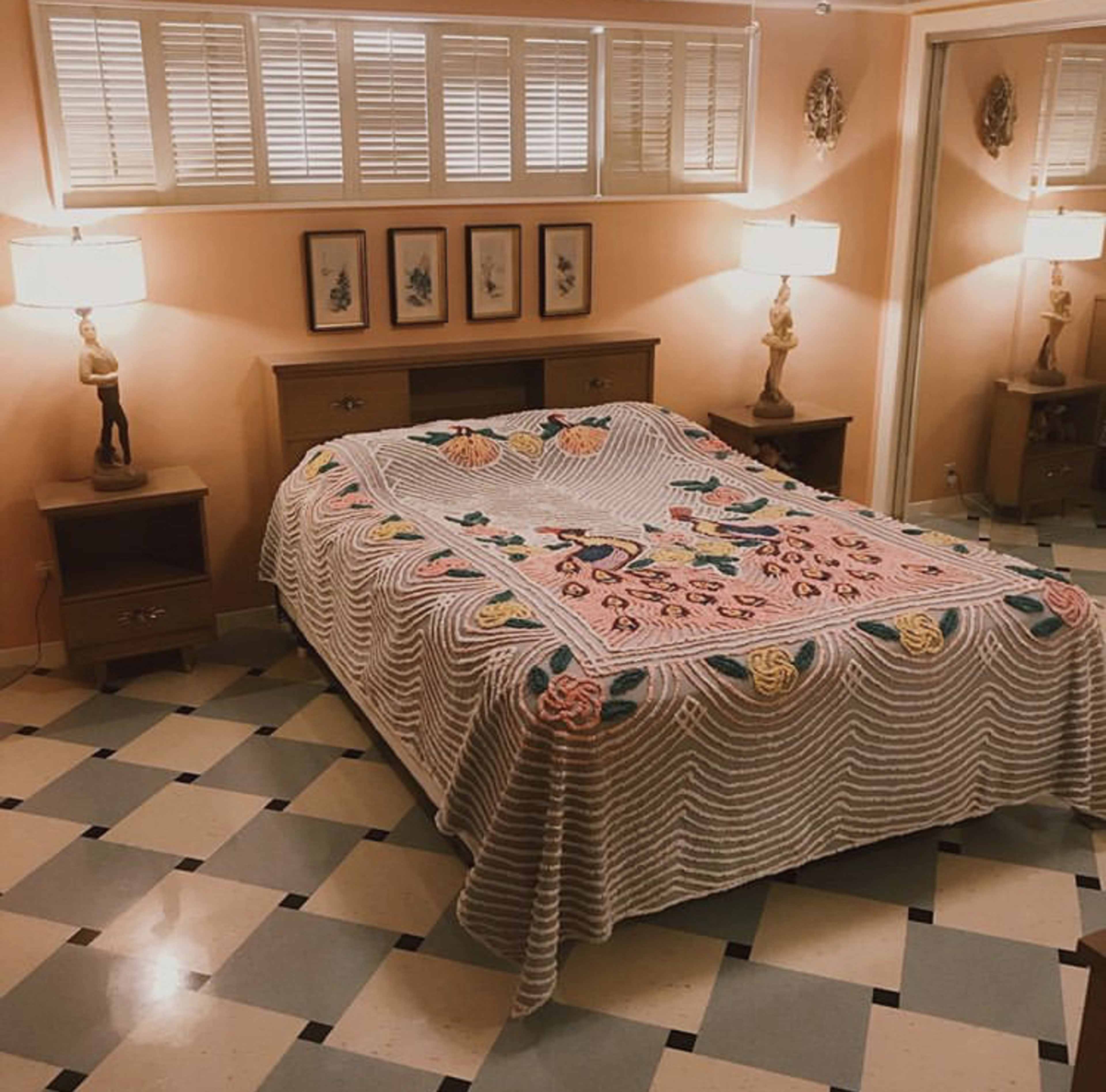 A bedroom features a bed with a colorful quilt, two bedside lamps, and framed artwork on the wall, set against a patterned tile floor.