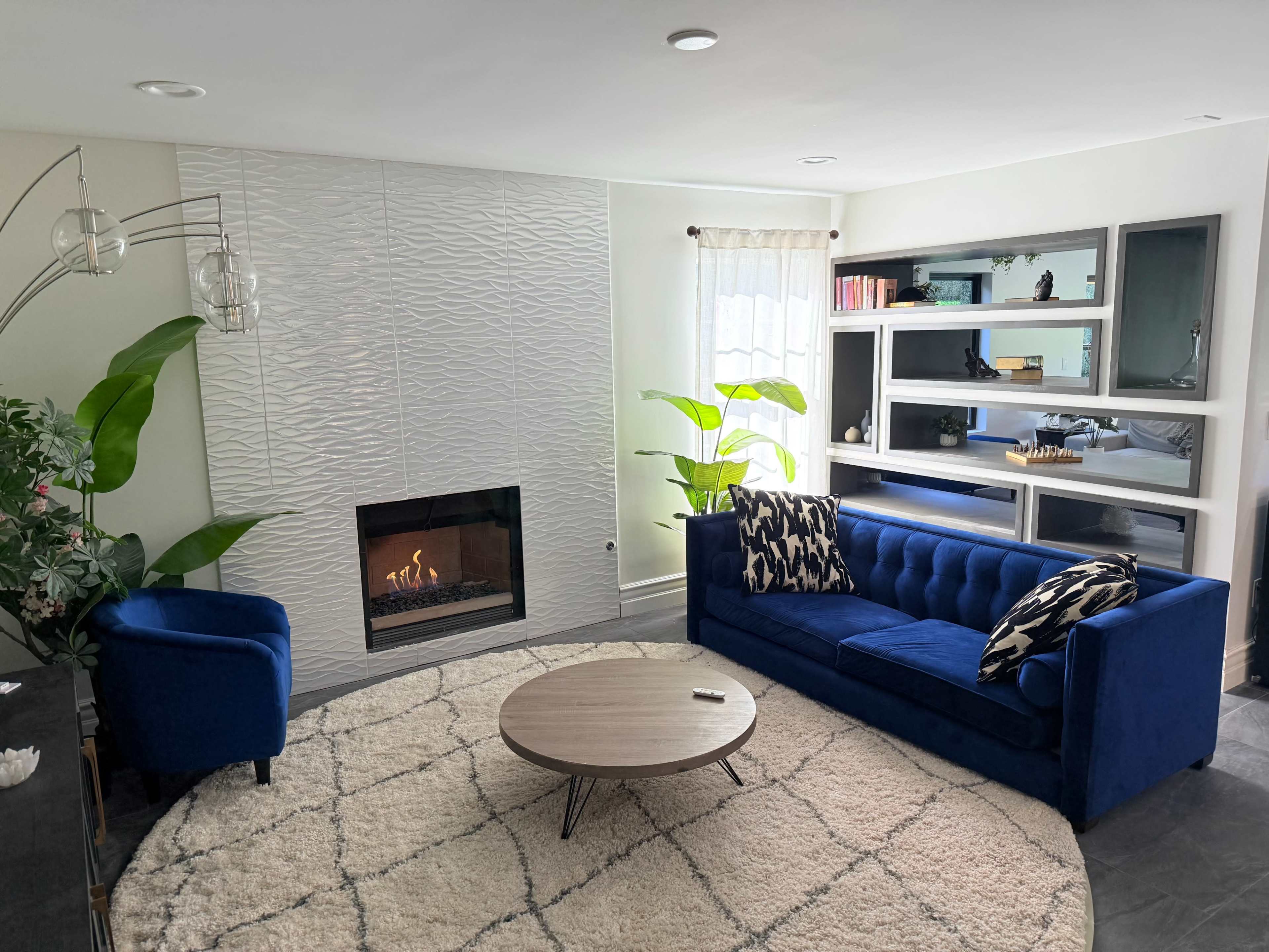 The image shows a modern living room featuring a blue velvet sofa and armchair, a round coffee table, a textured fireplace, and shelves with decorative items.