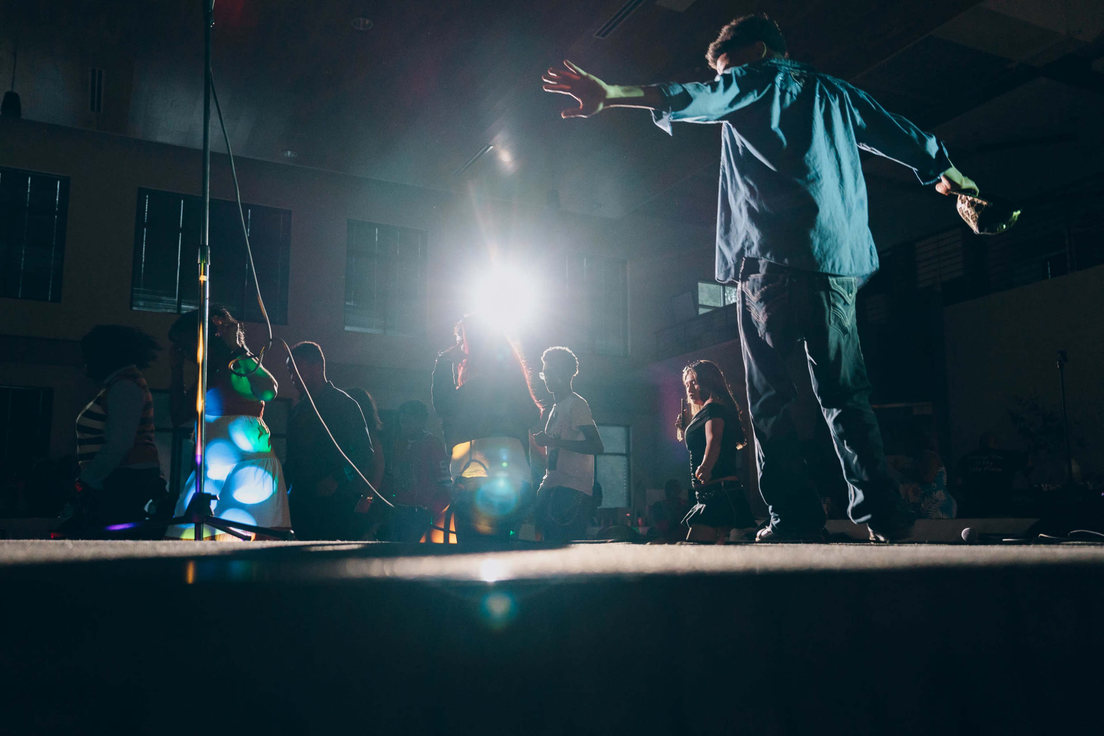 A performer gestures to an audience in a dimly lit venue, with a spotlight illuminating the scene.