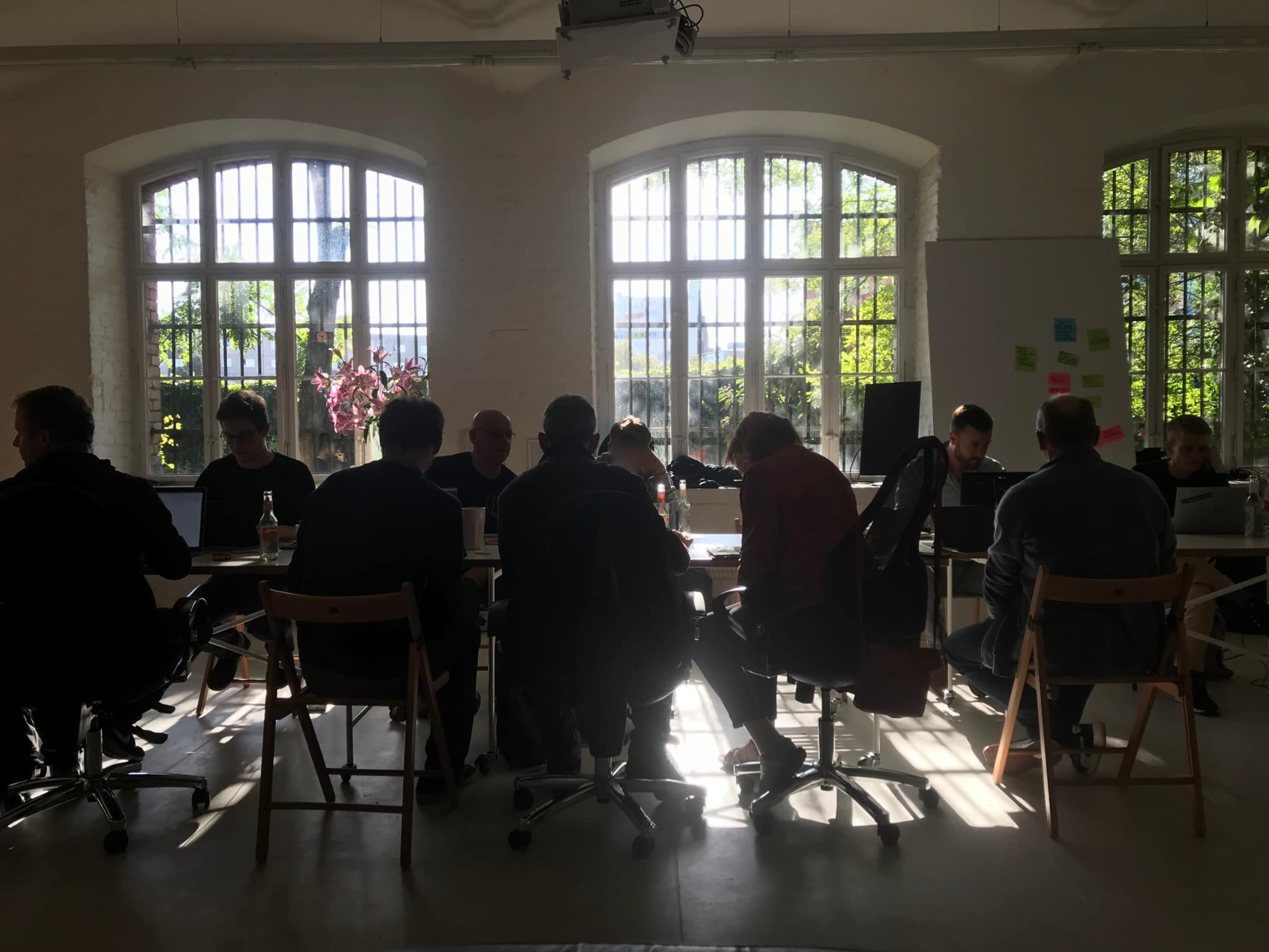 A group of people work around a table in a well-lit room with large windows, surrounded by plants.