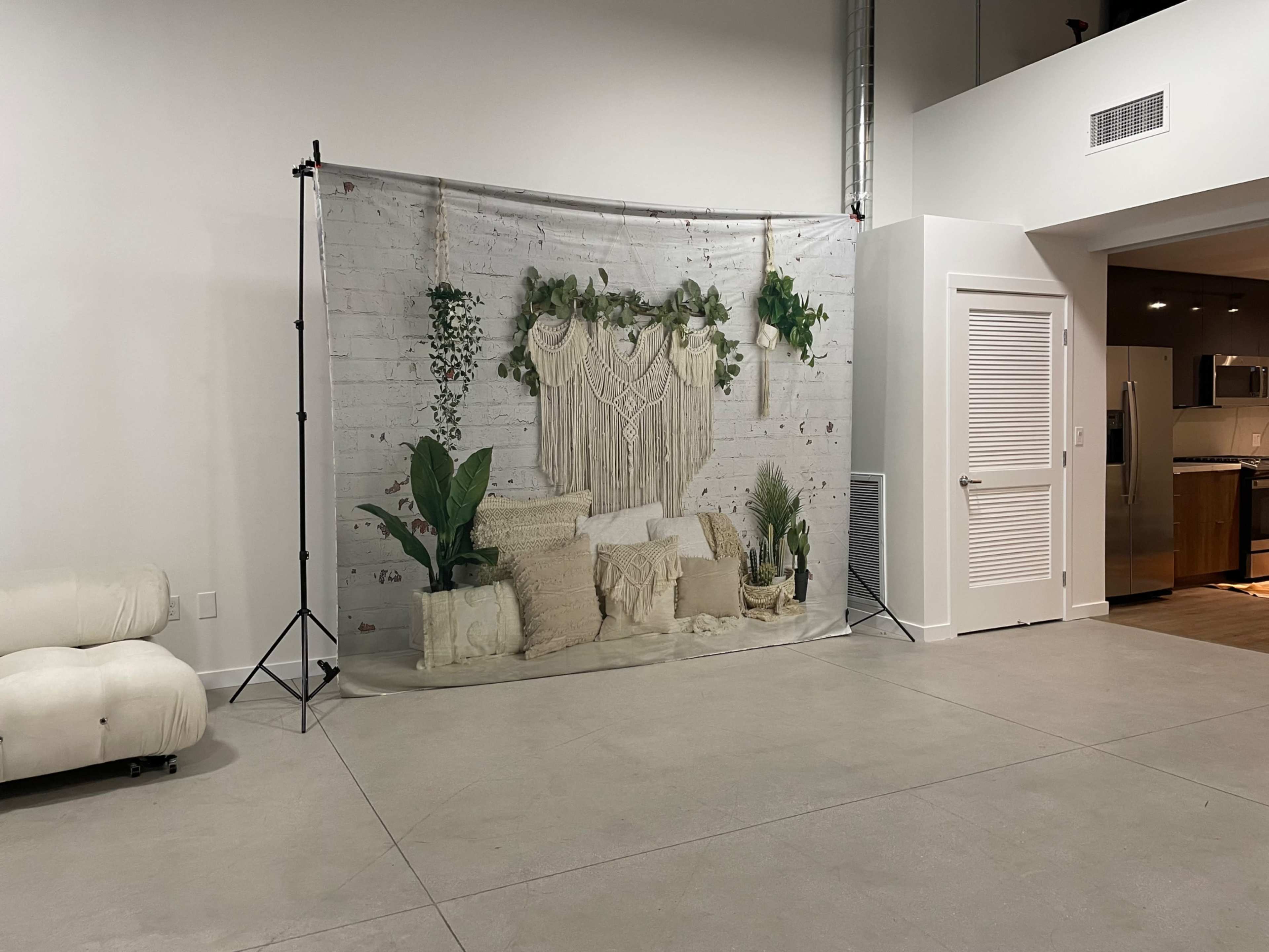 A decorative backdrop featuring a macramé wall hanging and various plants is positioned in a spacious, minimalist room with concrete flooring.