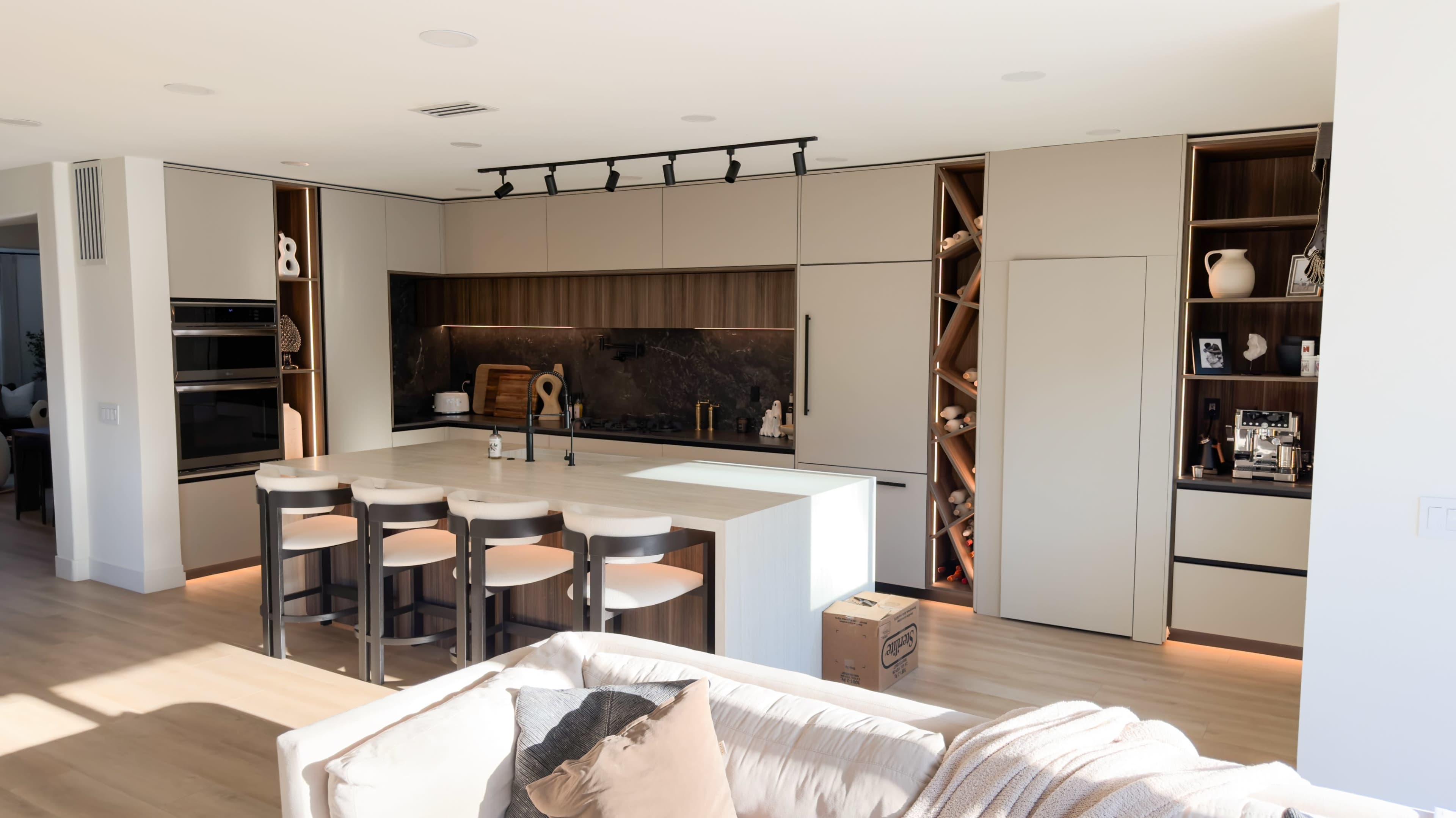 The image shows a modern kitchen with a central island, bar stools, sleek cabinets, and minimalistic decor, featuring a mix of wood and stone elements.