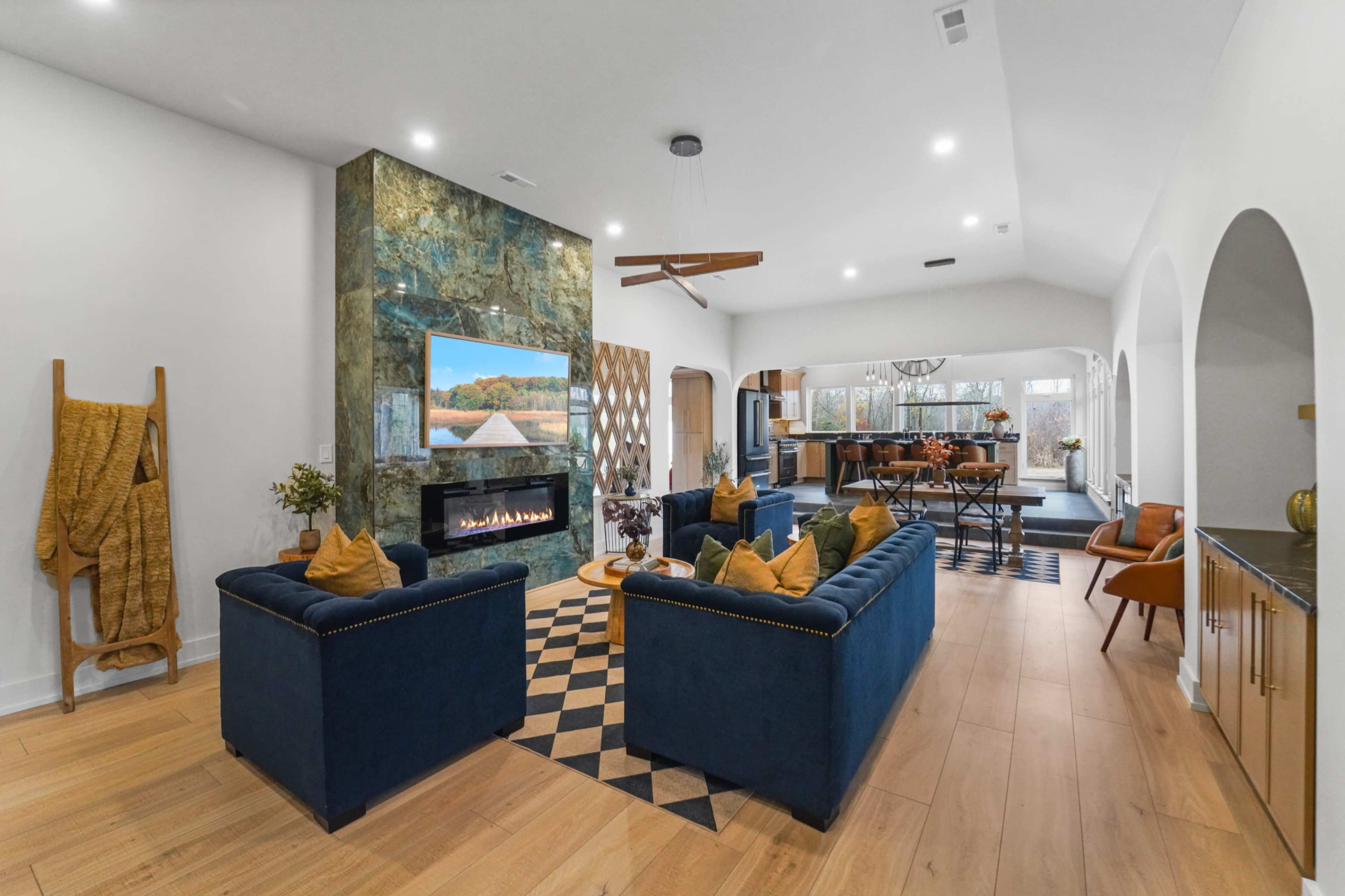 The image shows a modern living room featuring a blue sofa set arranged around a central fireplace with a large television above it, illuminated by natural light from large windows.