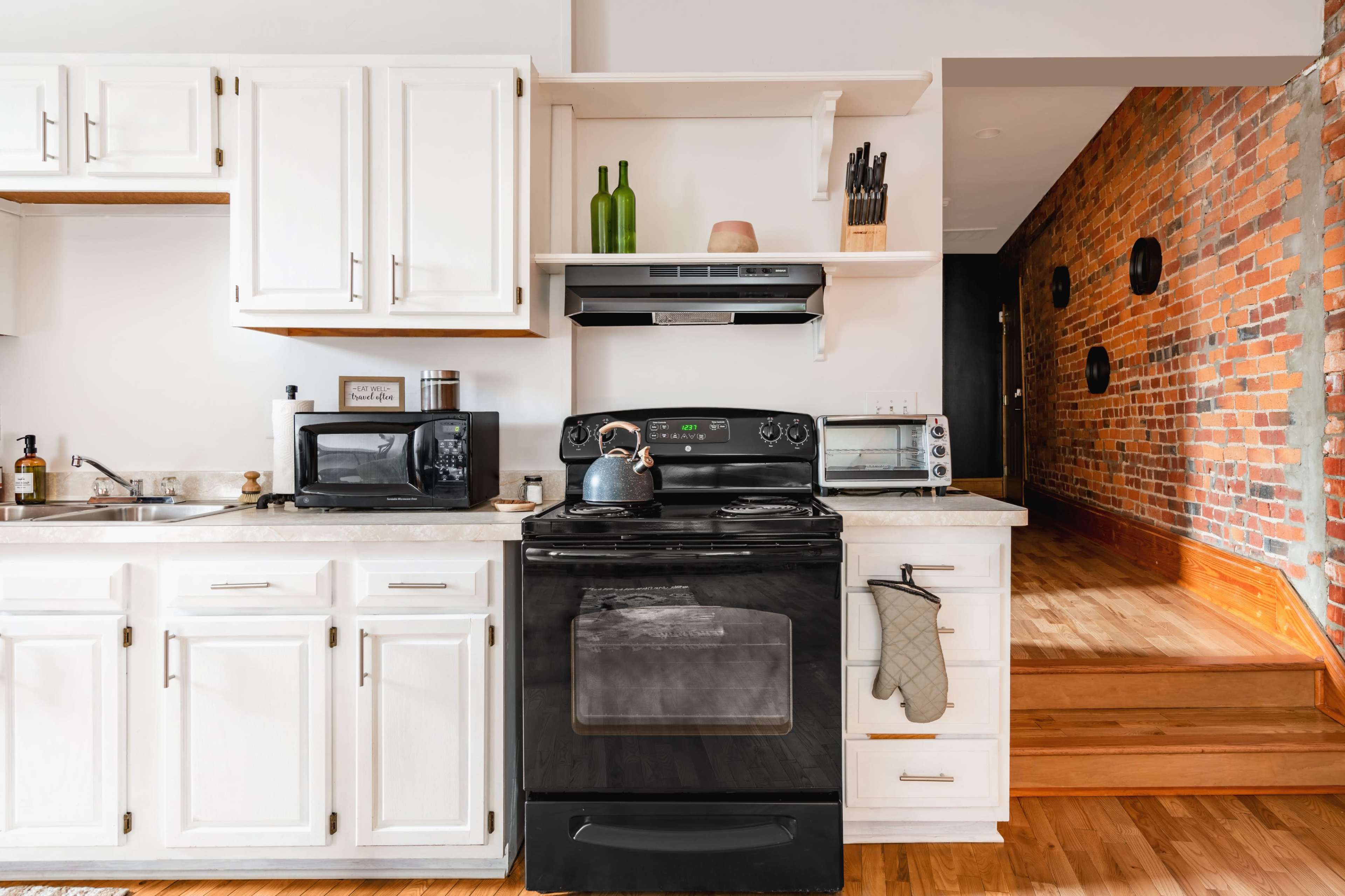 A modern kitchen features white cabinets, a black stove, a microwave, and wooden flooring, with a staircase leading to a room adorned with brick walls.