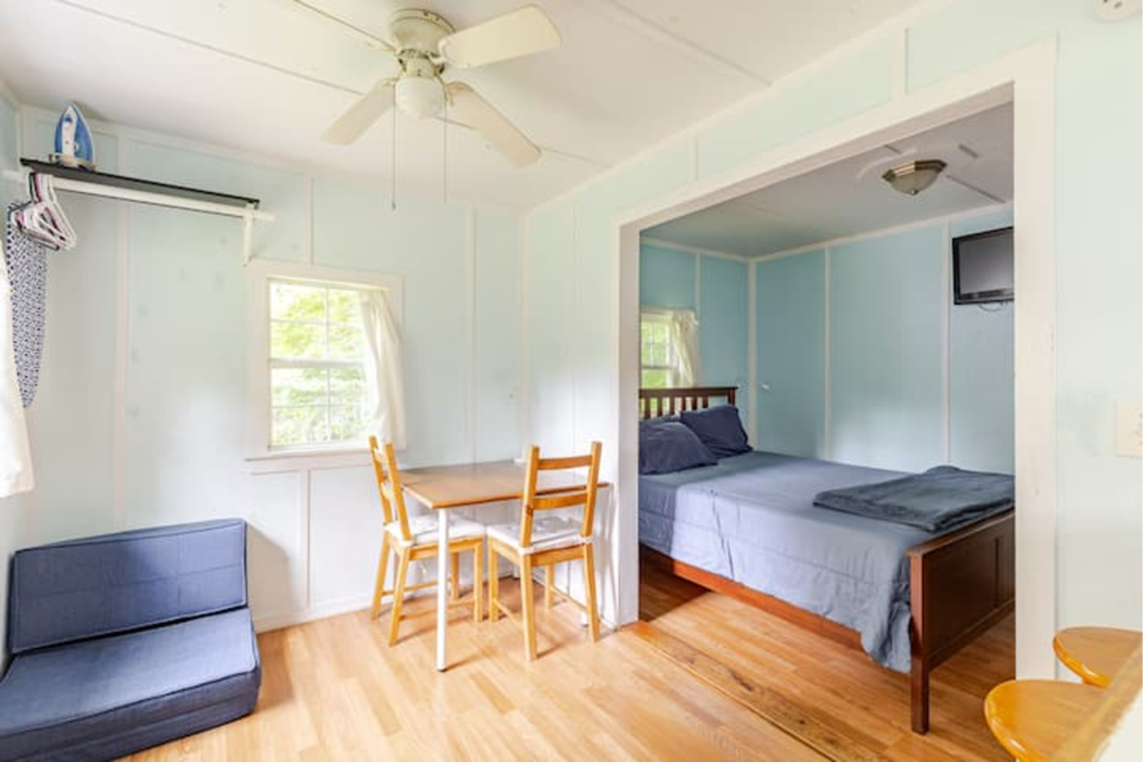 A small, light blue room features a double bed, a table with two chairs, a small couch, and a ceiling fan.