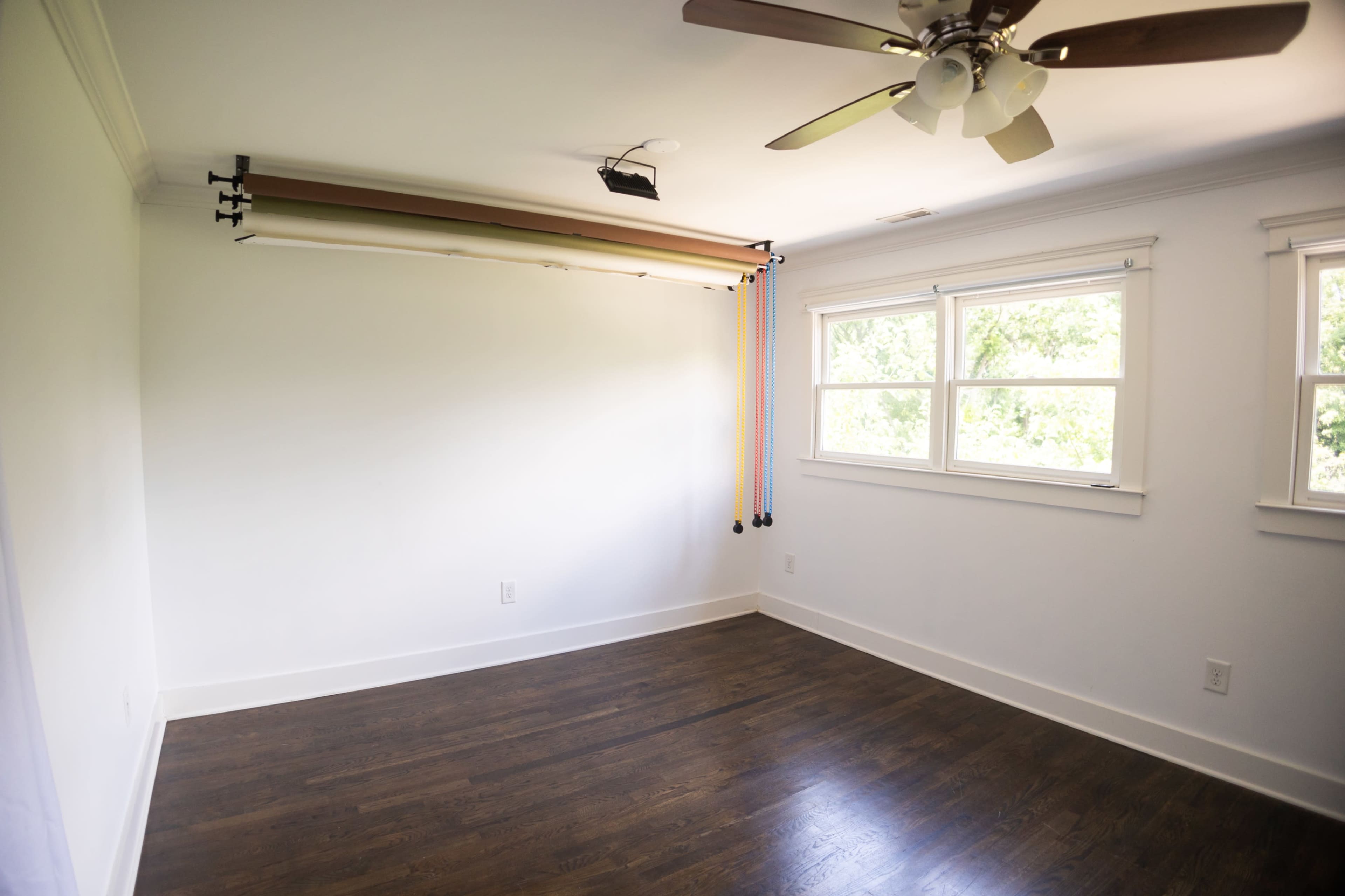 A bright, empty room with a wooden floor features a ceiling fan, two windows, and a roll-up backdrop system mounted on the wall.