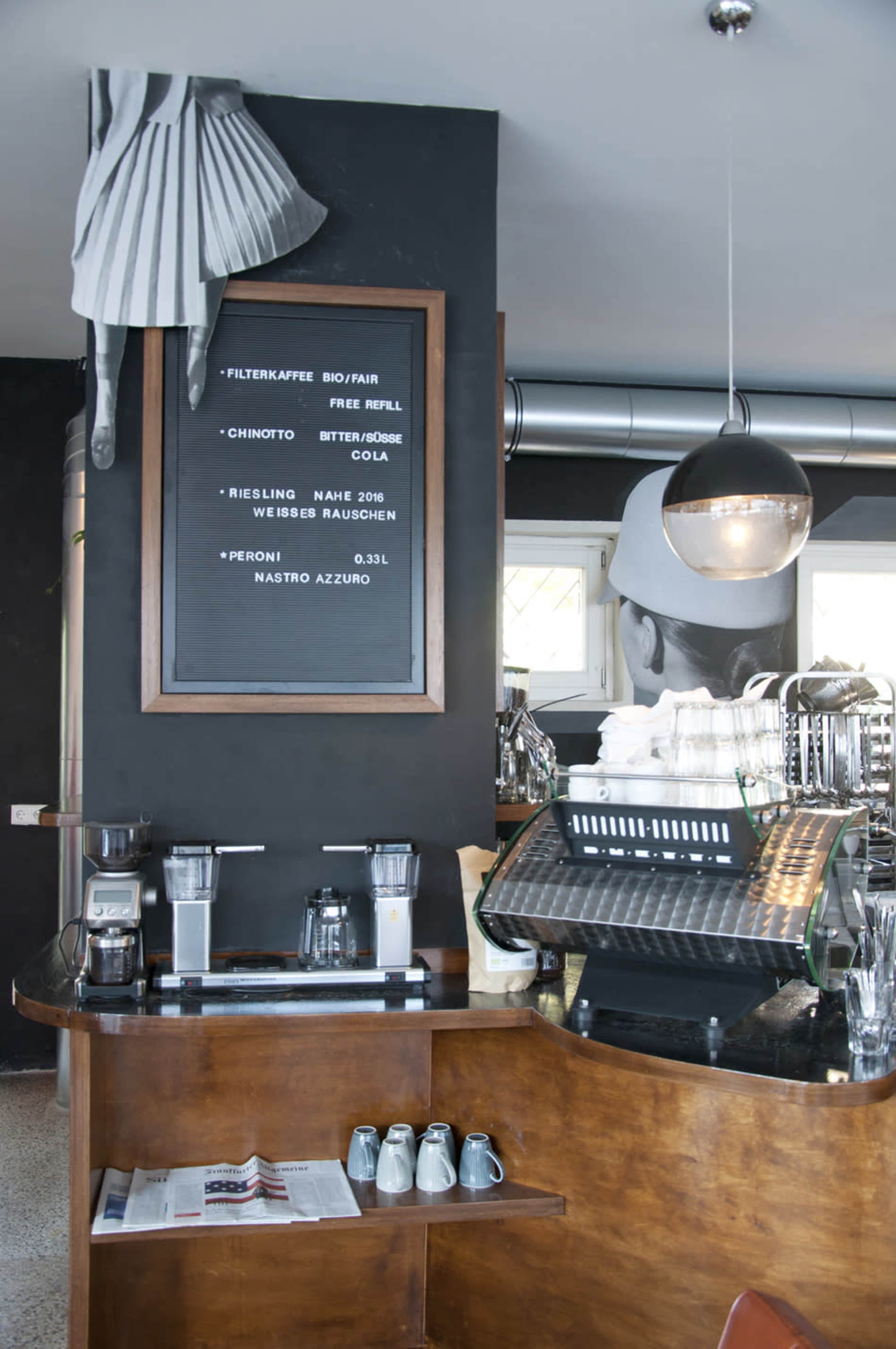 The image shows a modern coffee bar with a dark wall featuring a chalkboard menu, metallic accents, and a curved wooden counter with coffee equipment and glasses displayed.