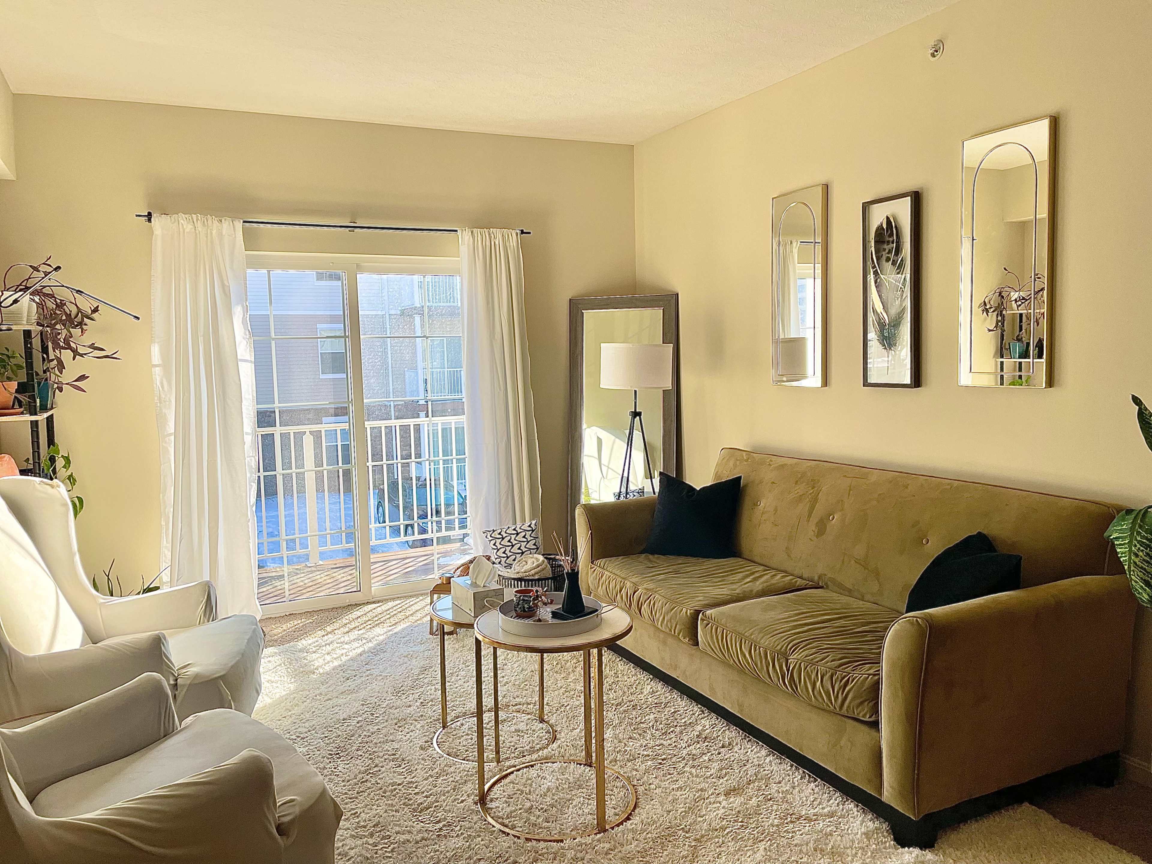 A cozy living room with a green sofa, two side tables, and large windows revealing a balcony.