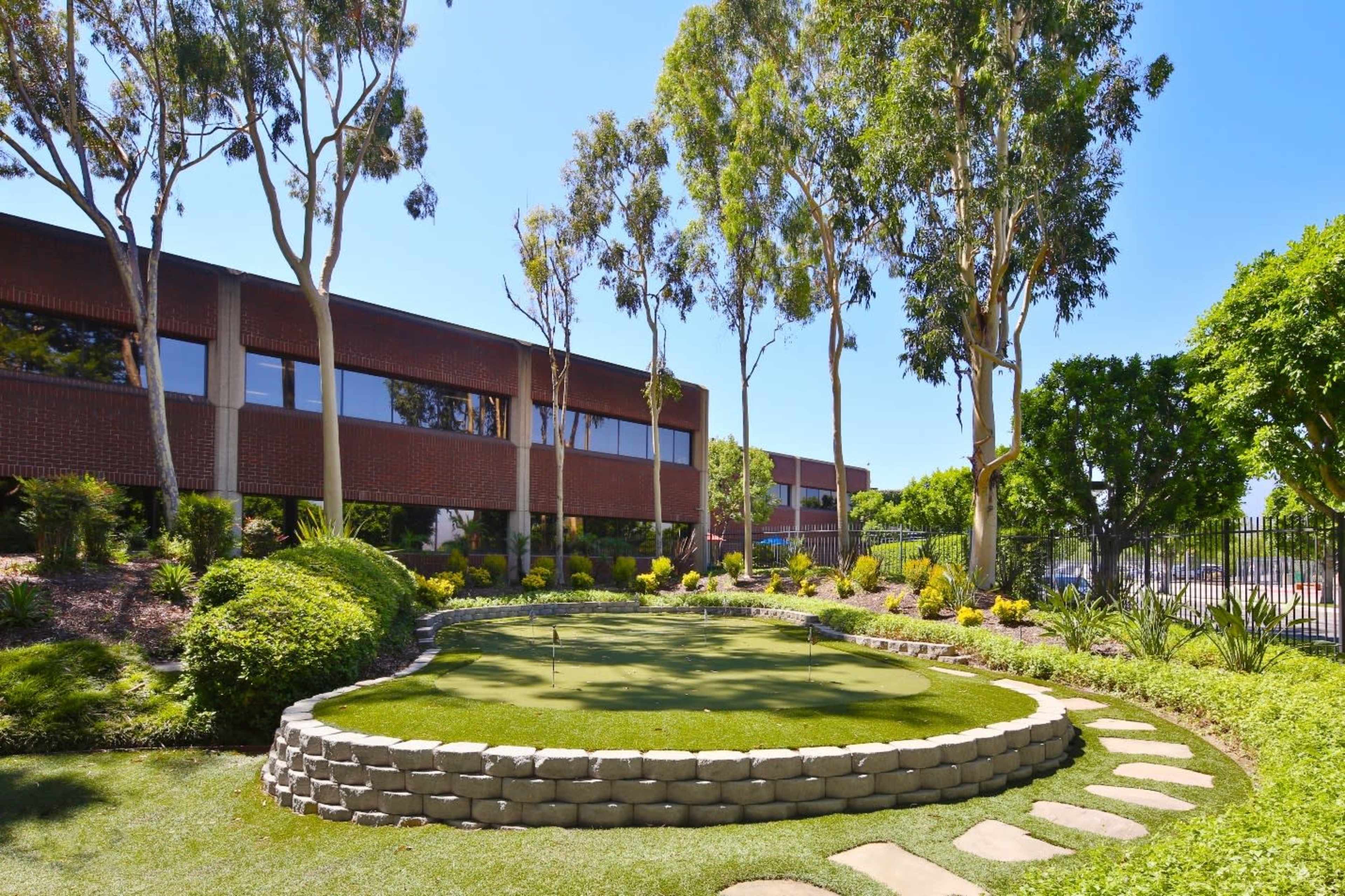 A landscaped garden with a small putting green bordered by stone, palm trees, and a brick building in the background.