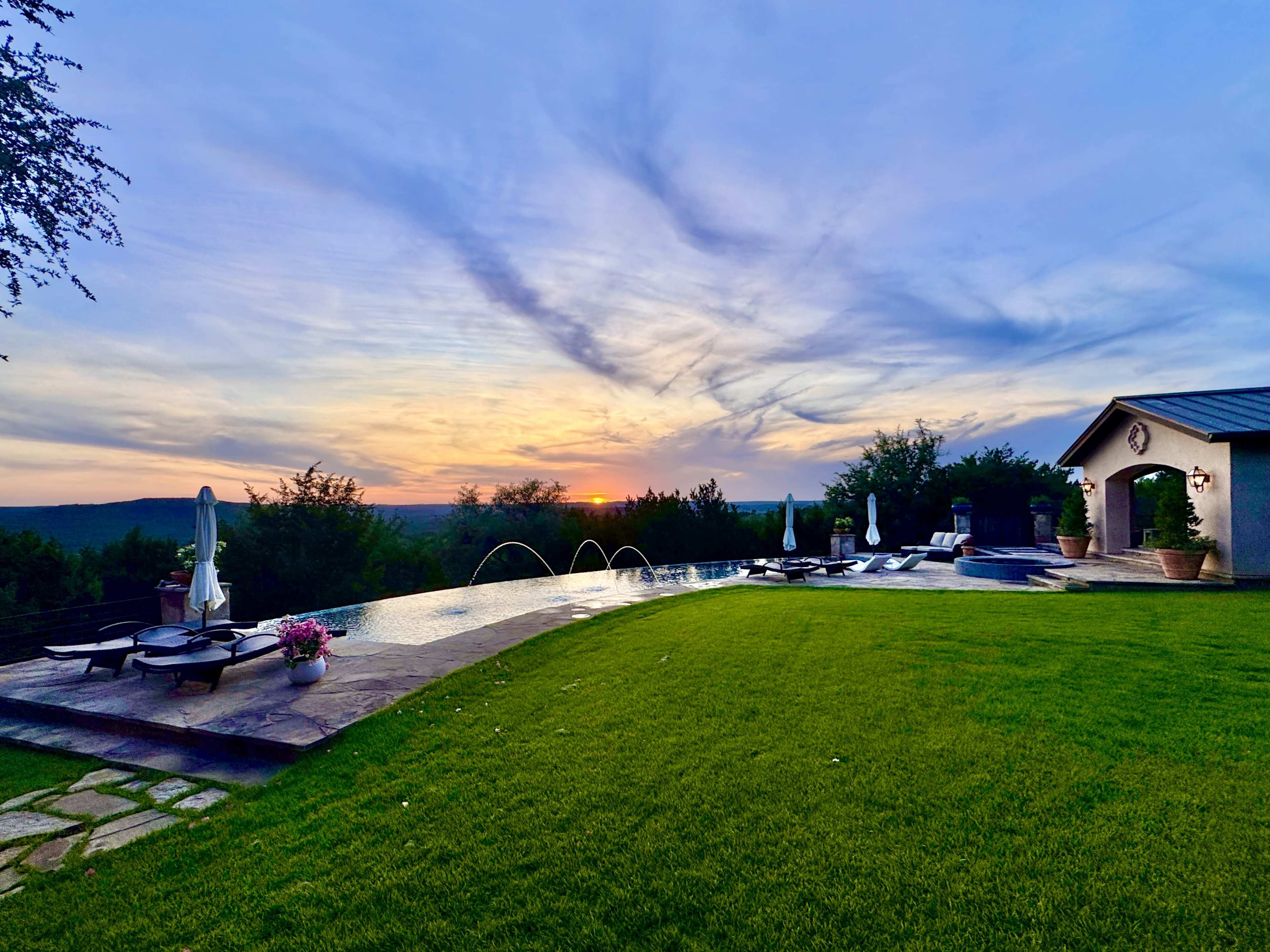 A luxurious backyard features a spacious lawn, an infinity pool with fountains, and a sunset view over distant hills.