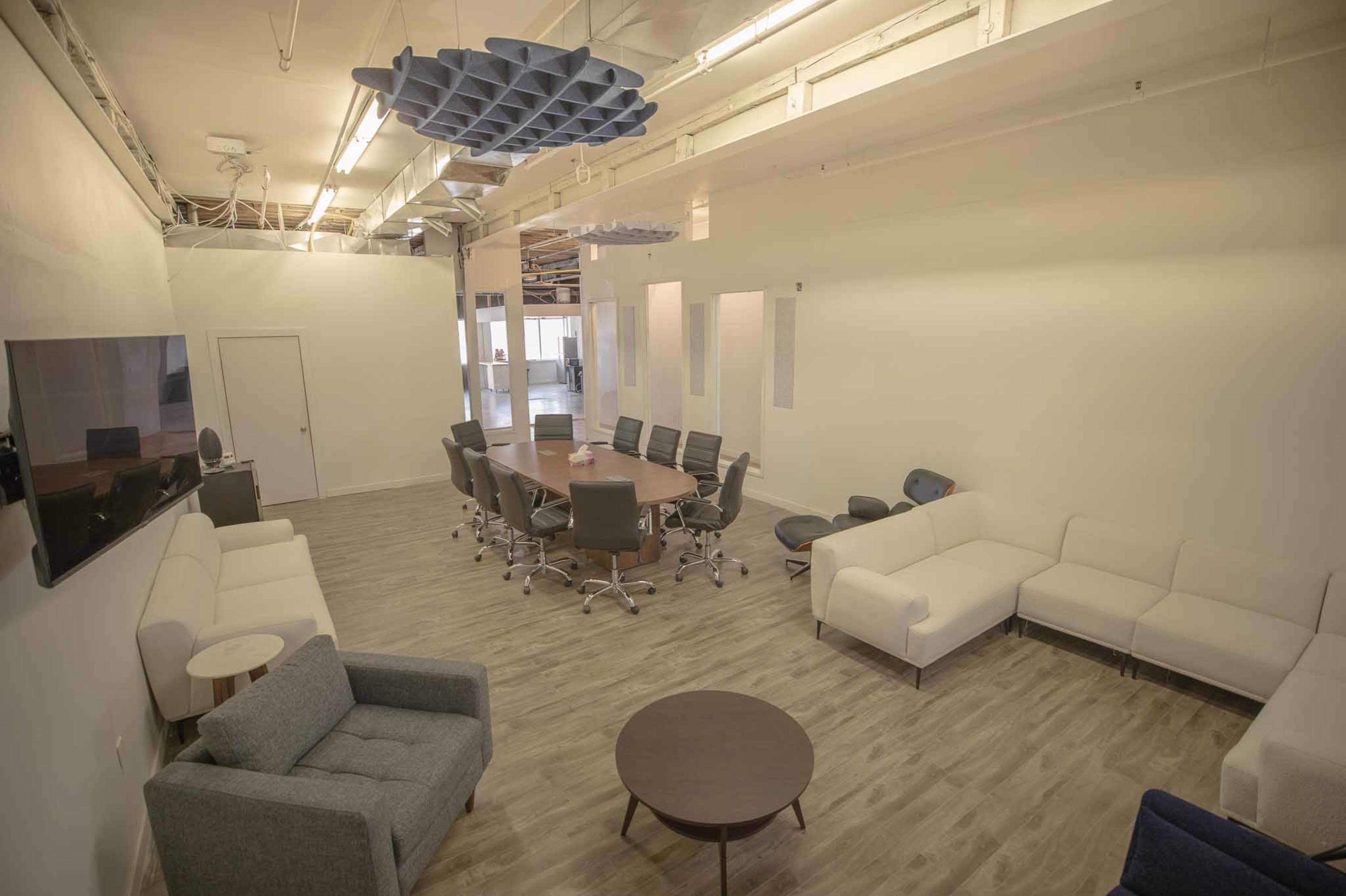 The image shows a modern office space featuring a large conference table surrounded by chairs, a seating area with couches, and a circular coffee table, all under a ceiling with unique acoustic panels.