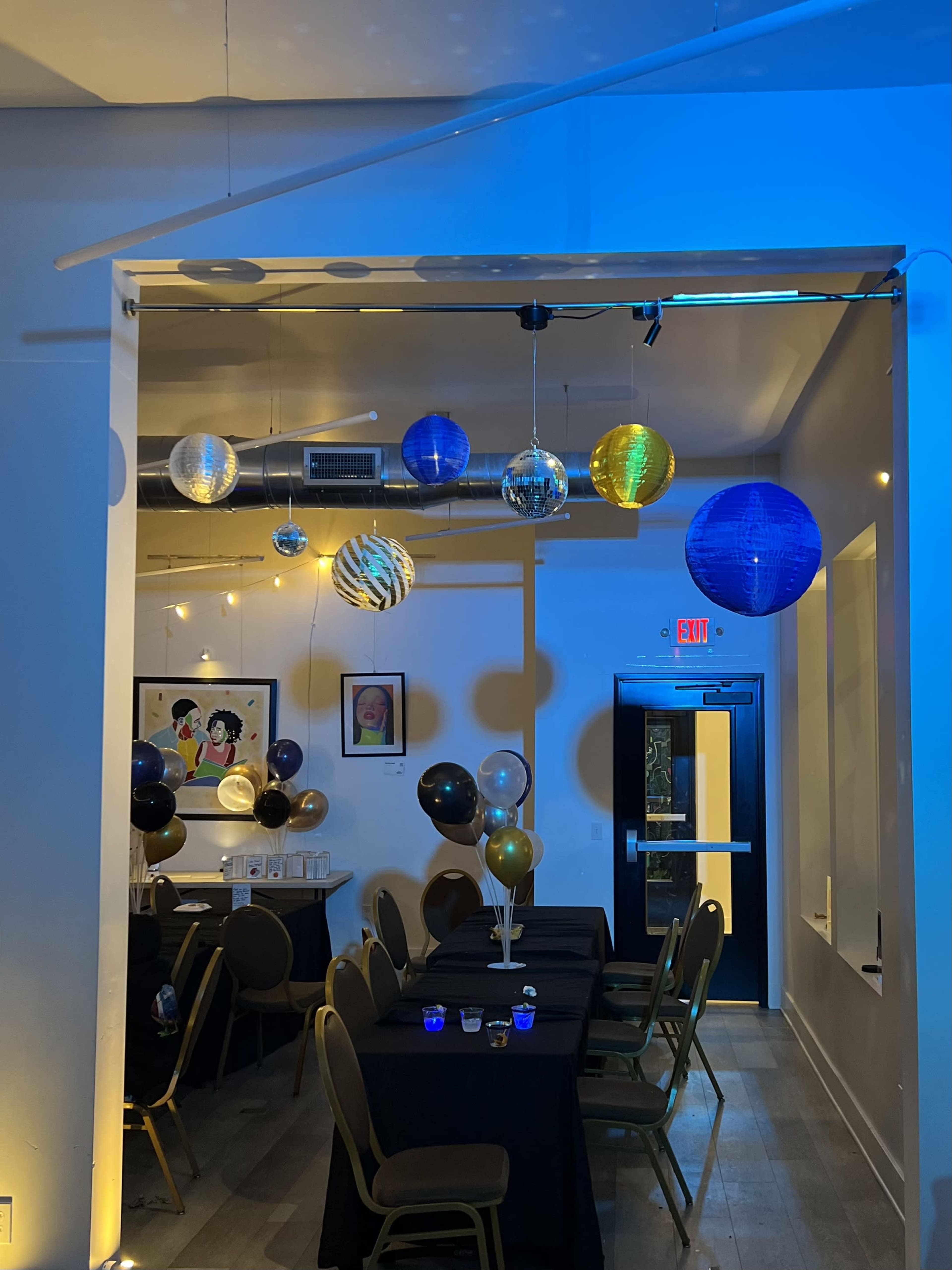 The image shows a decorated indoor event space with tables covered in black cloth, colorful hanging decorations, and balloons.