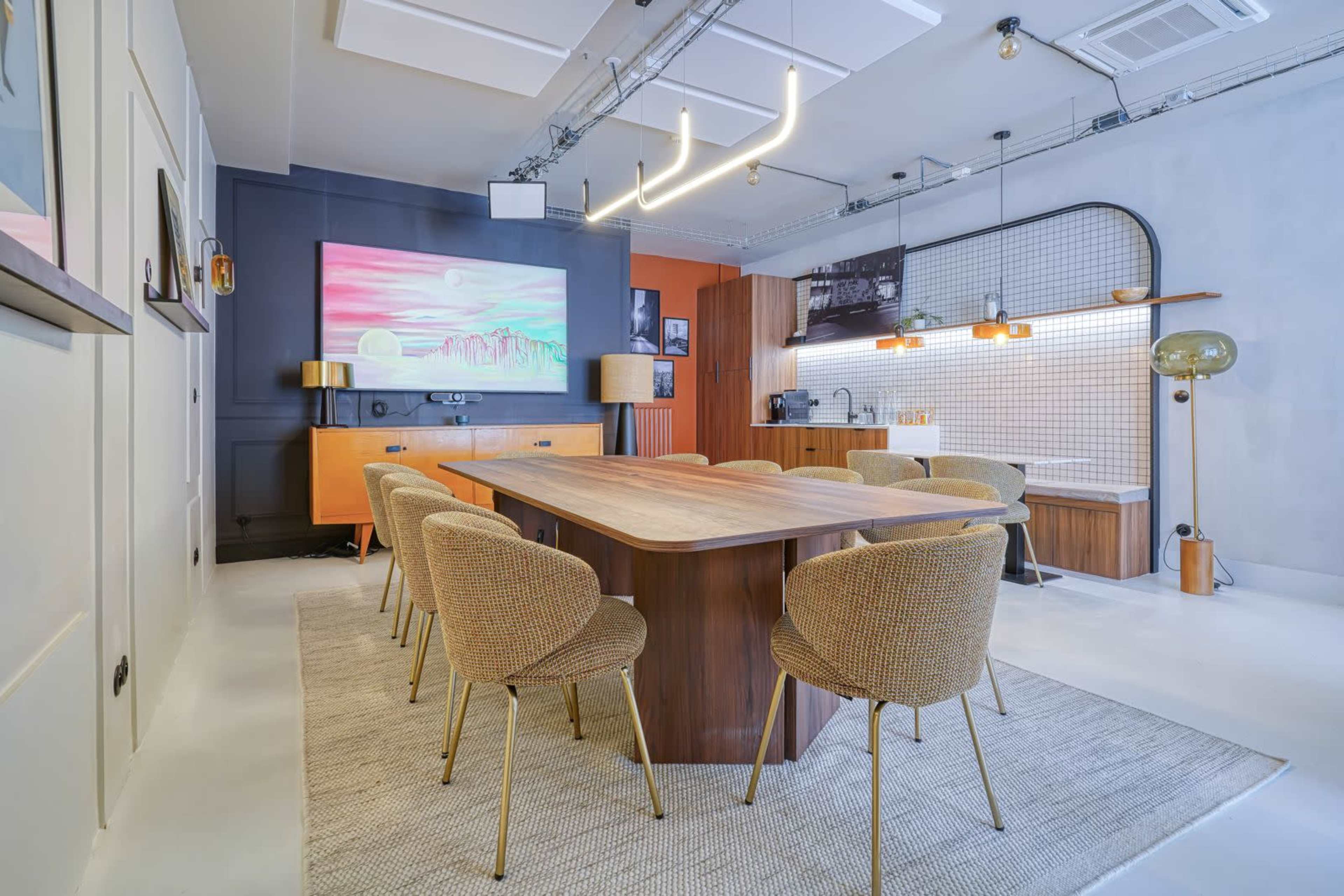 A modern dining area features a large wooden table surrounded by upholstered chairs, with a wall-mounted television and a kitchen area in the background.