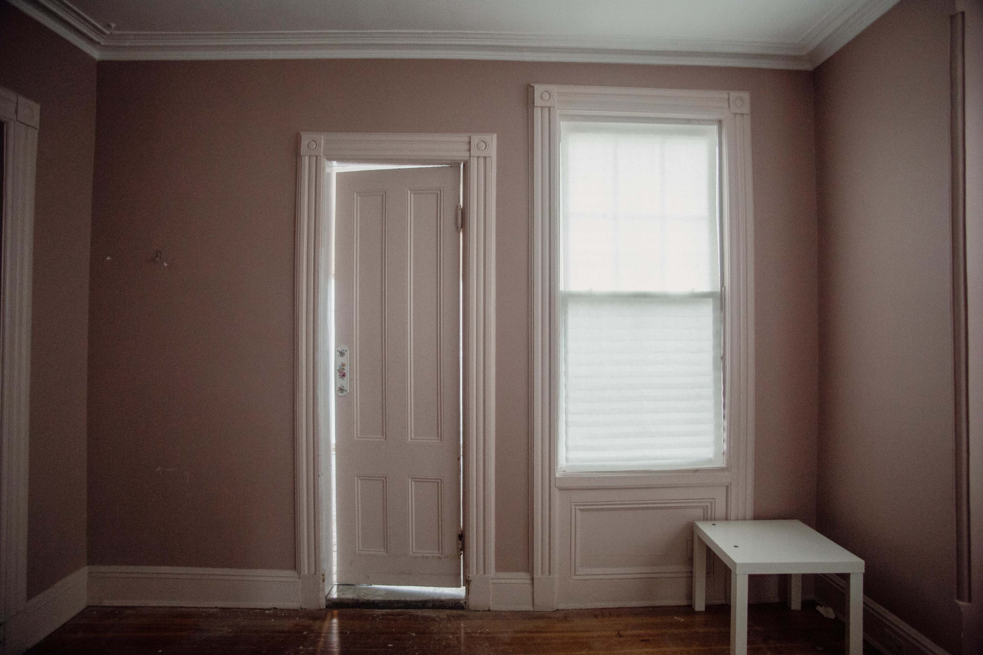 The image shows a minimalistic room with light pink walls, a partially open door, a window with blinds, and a small white table.
