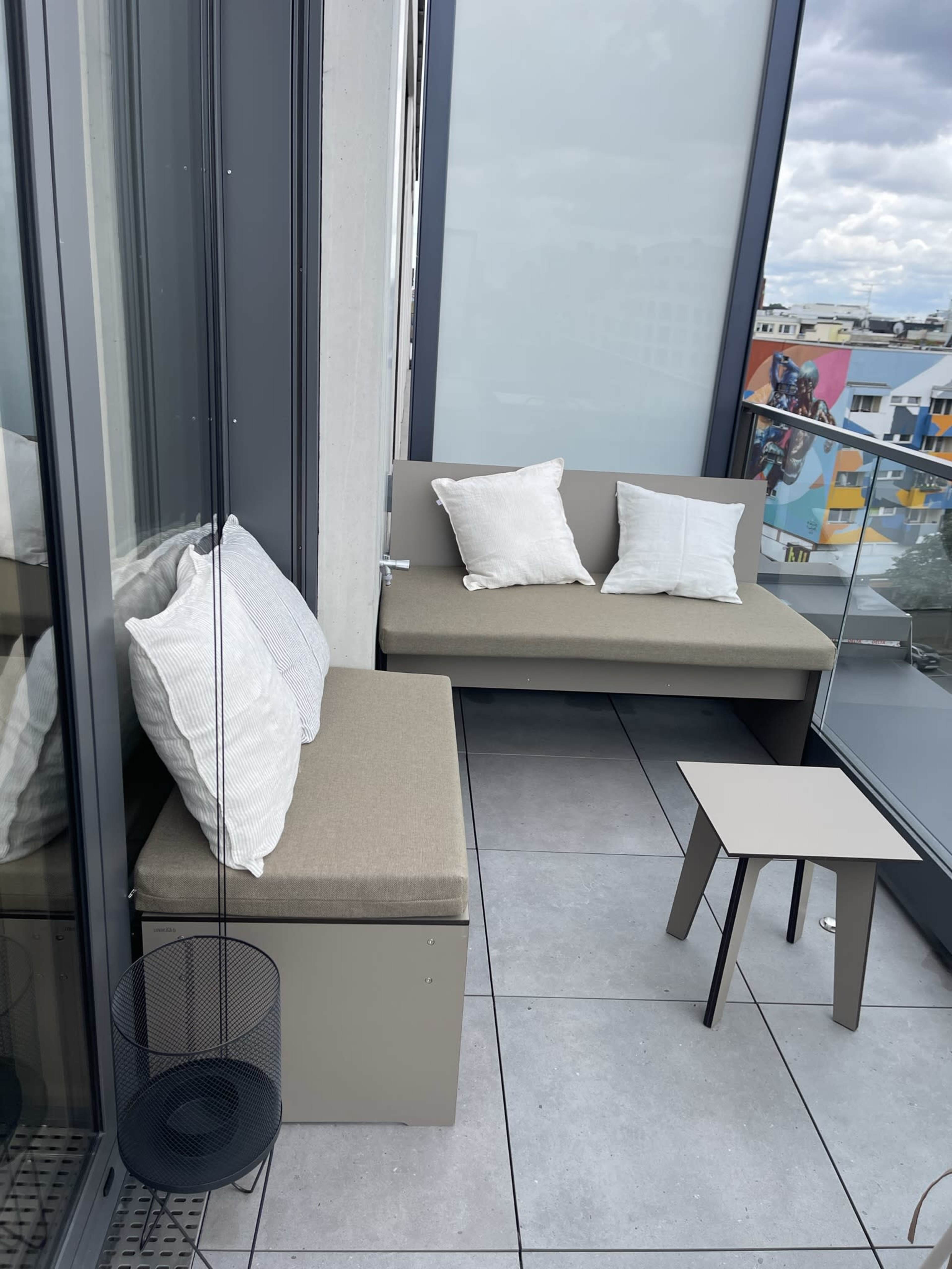 A balcony features a small seating area with a bench and two cushions, a side table, and a decorative planter.