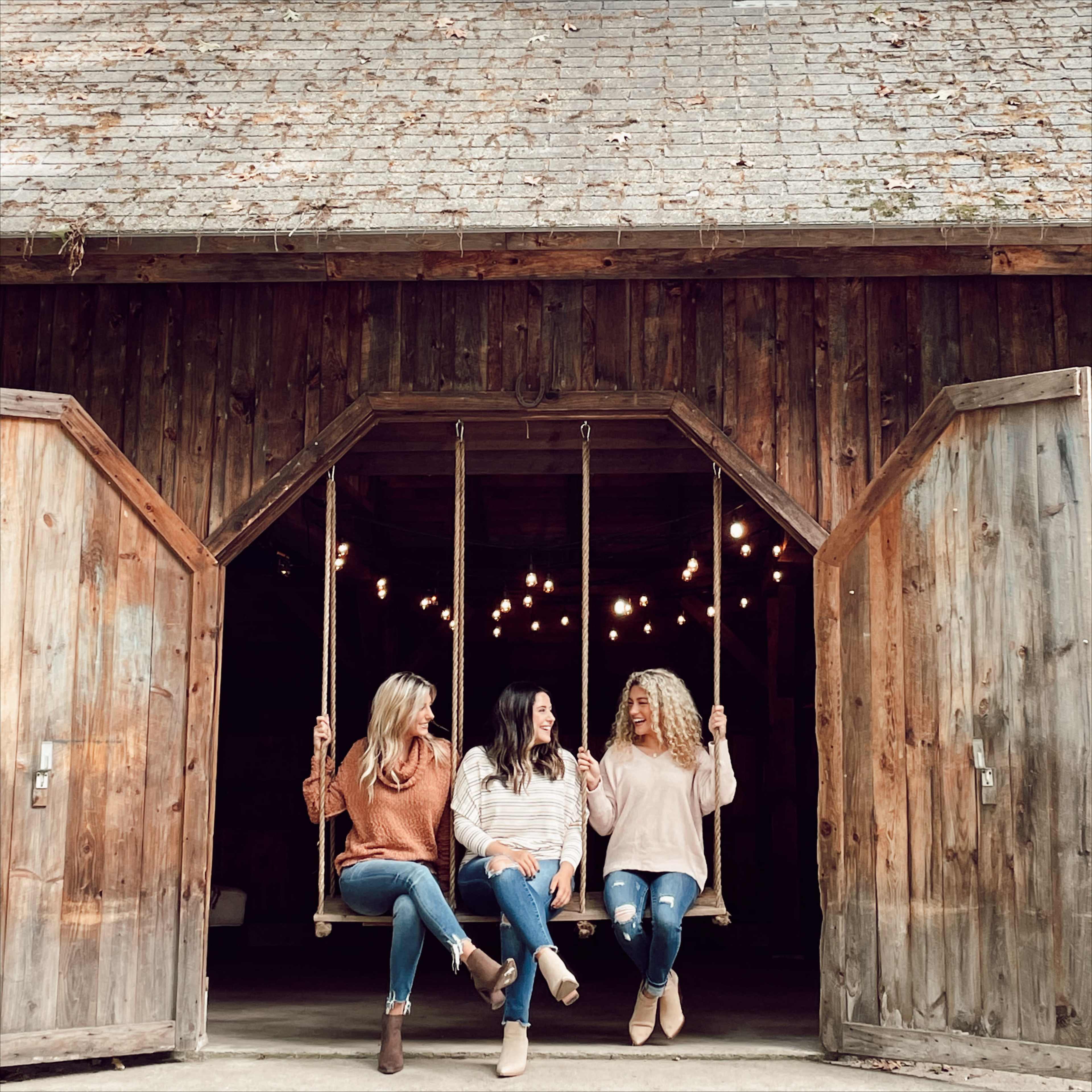 Three women sit on swings outside a wooden barn, with lights hanging above them.