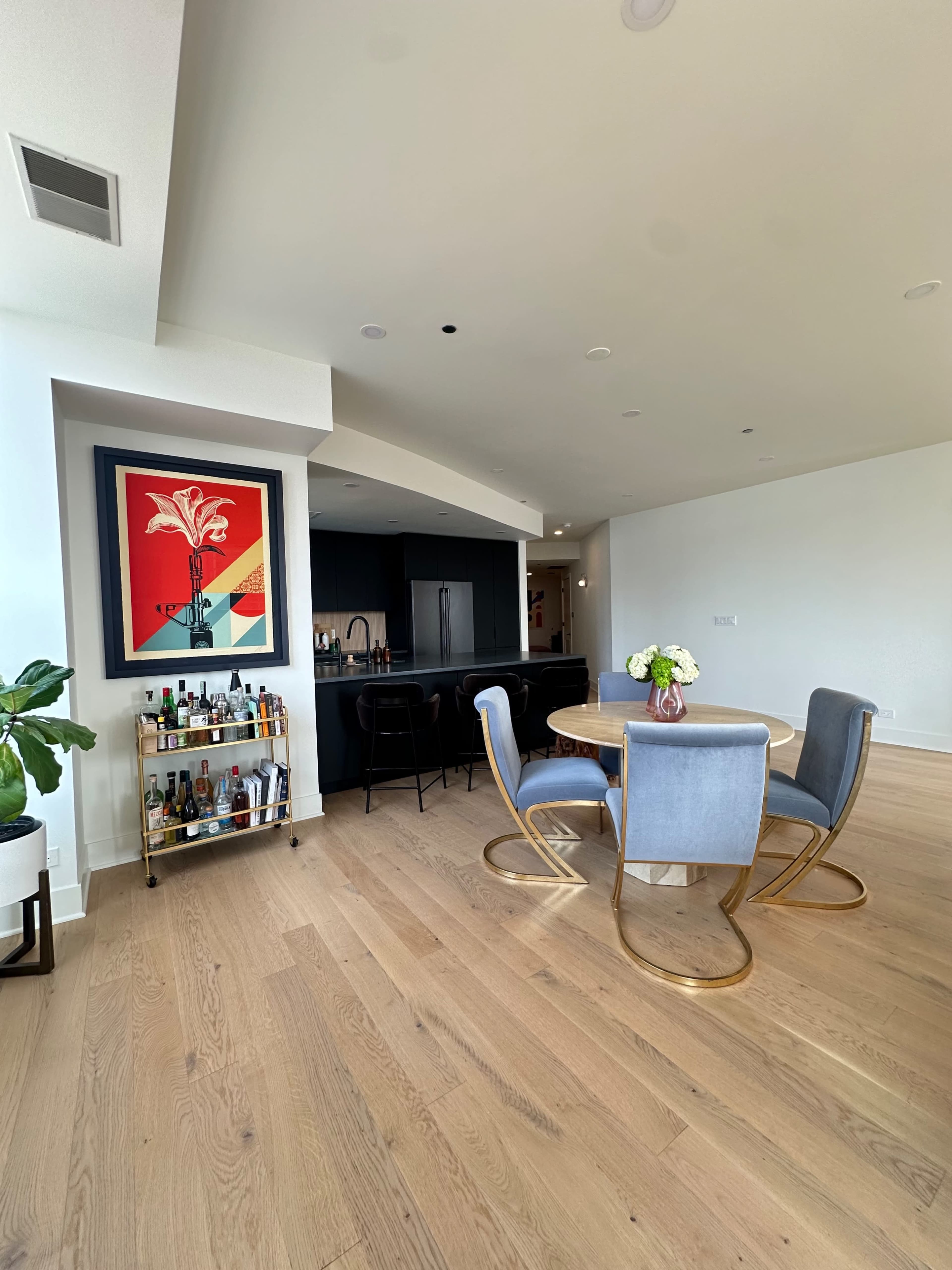 A modern dining area features a round table with four gray chairs, a bar cart filled with beverages, and a large artwork on the wall.