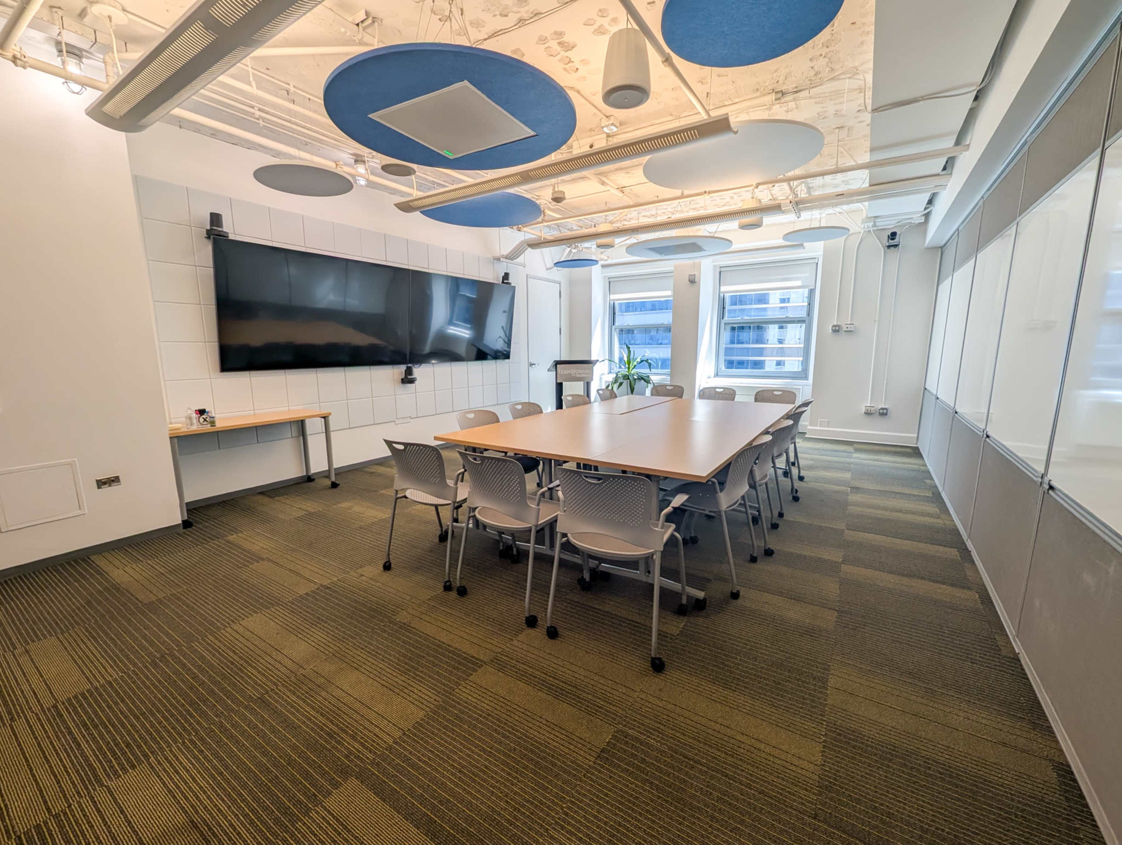 A modern conference room features a long table surrounded by chairs, with a large screen mounted on the wall and windows allowing natural light to enter.