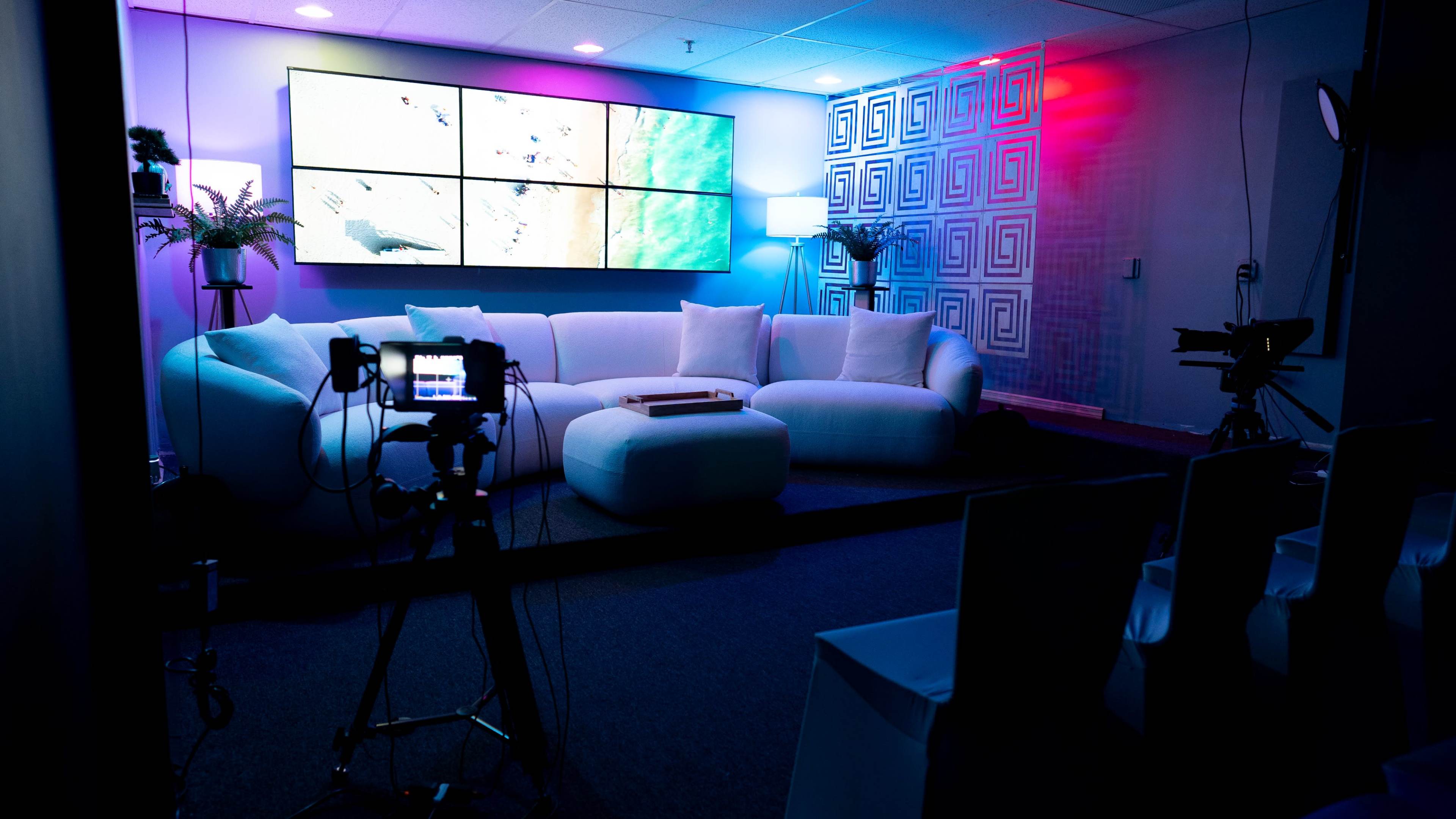 A modern studio setup featuring a white sectional sofa, a large display screen on the wall, and various lighting elements.