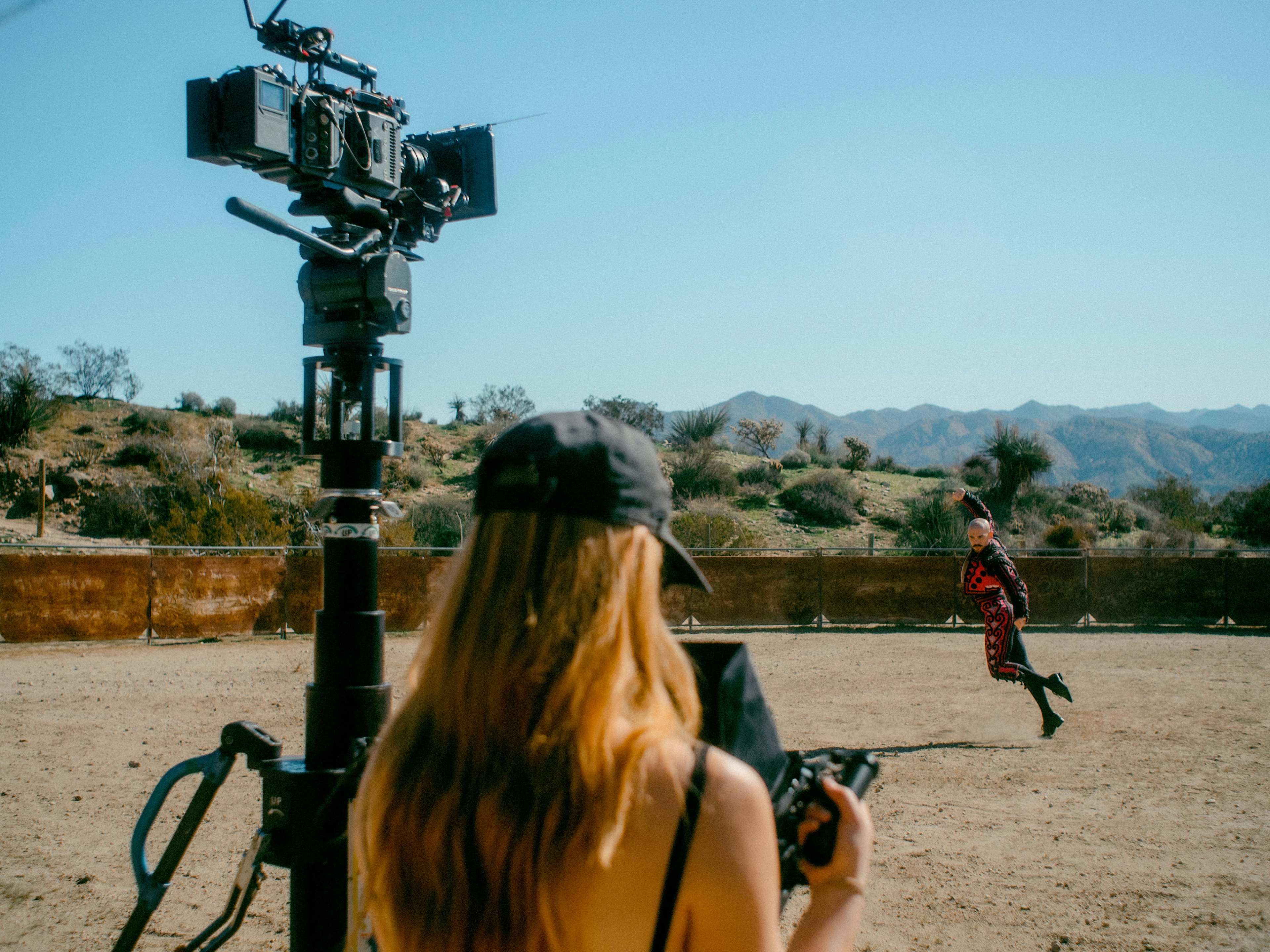 A camera operator captures a performer in a desert landscape as a film crew sets up equipment nearby.