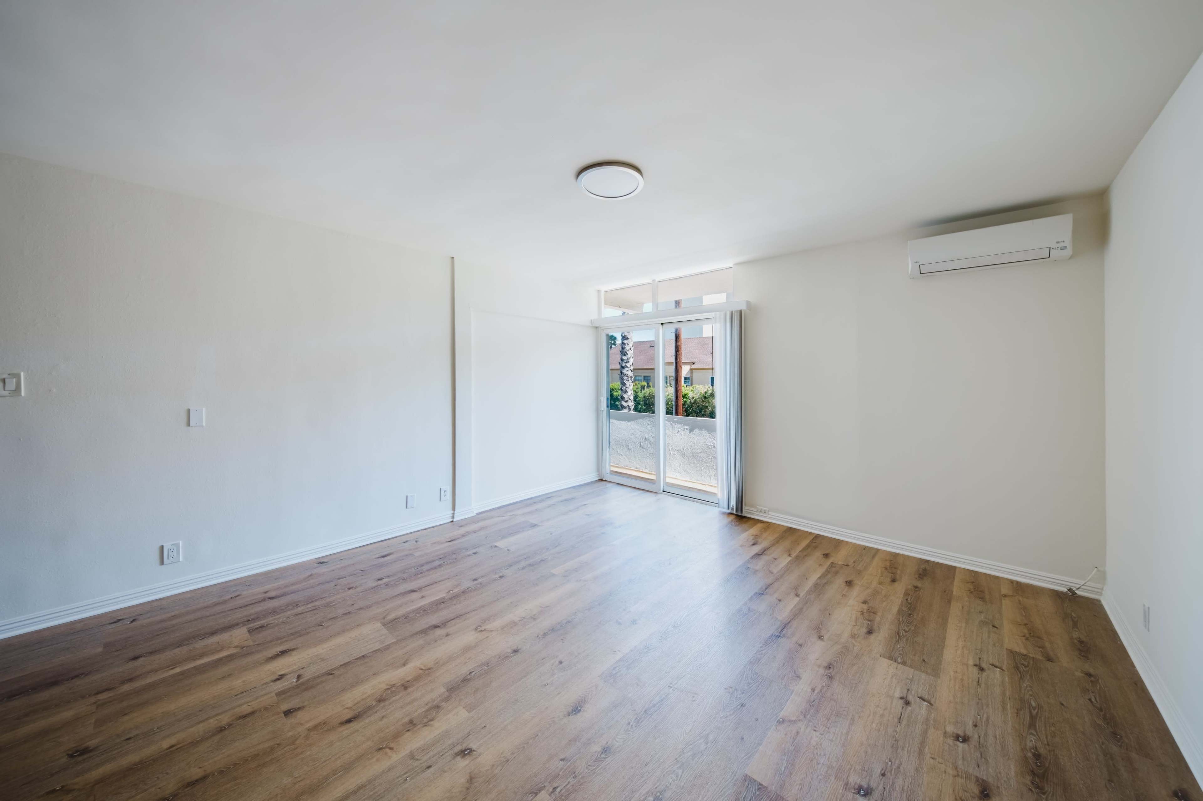 The image shows a spacious, unfurnished room with wooden flooring and a large sliding door leading to an outdoor area.