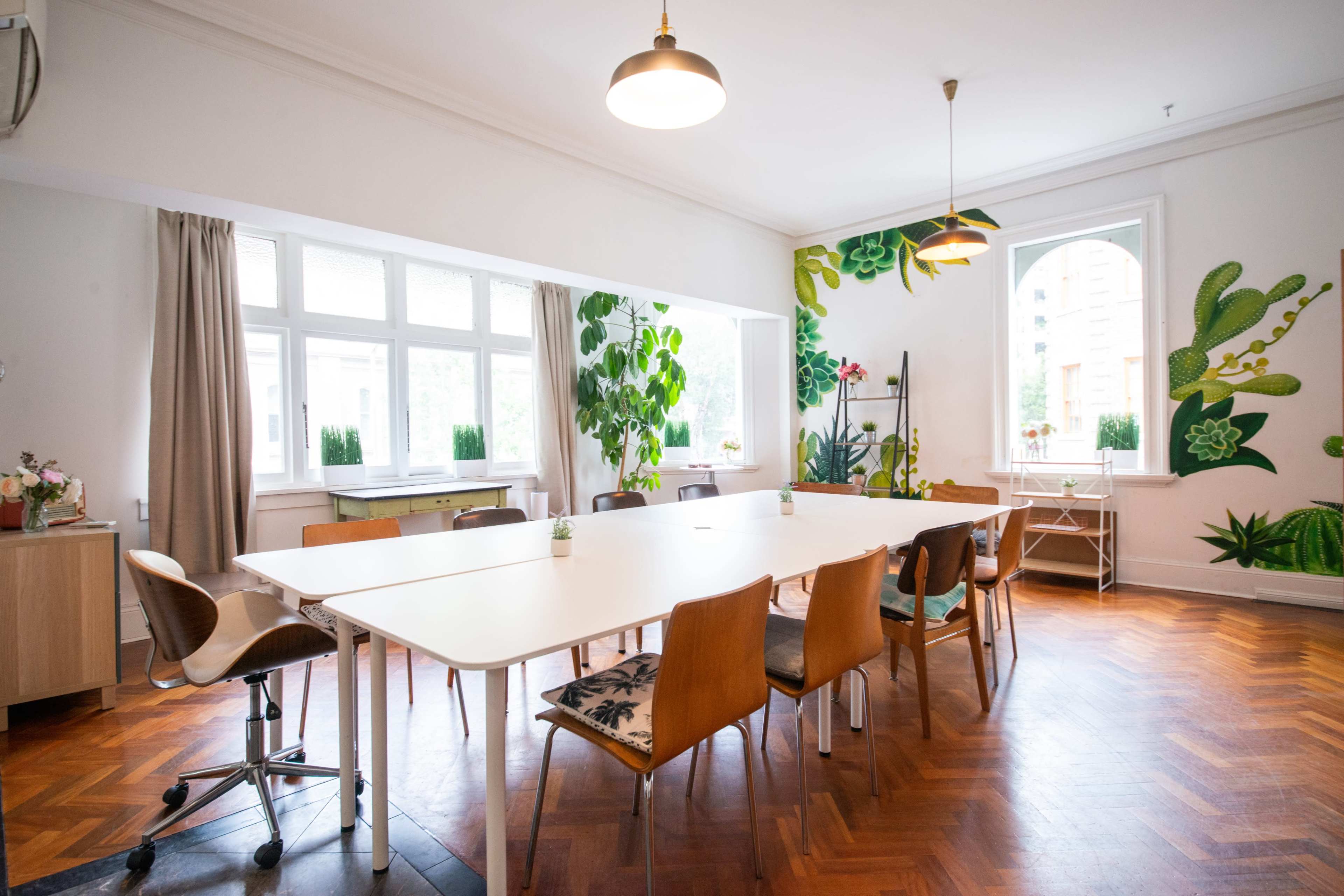 A bright meeting room features a large white table surrounded by wooden chairs and vibrant green plant decorations on the walls.