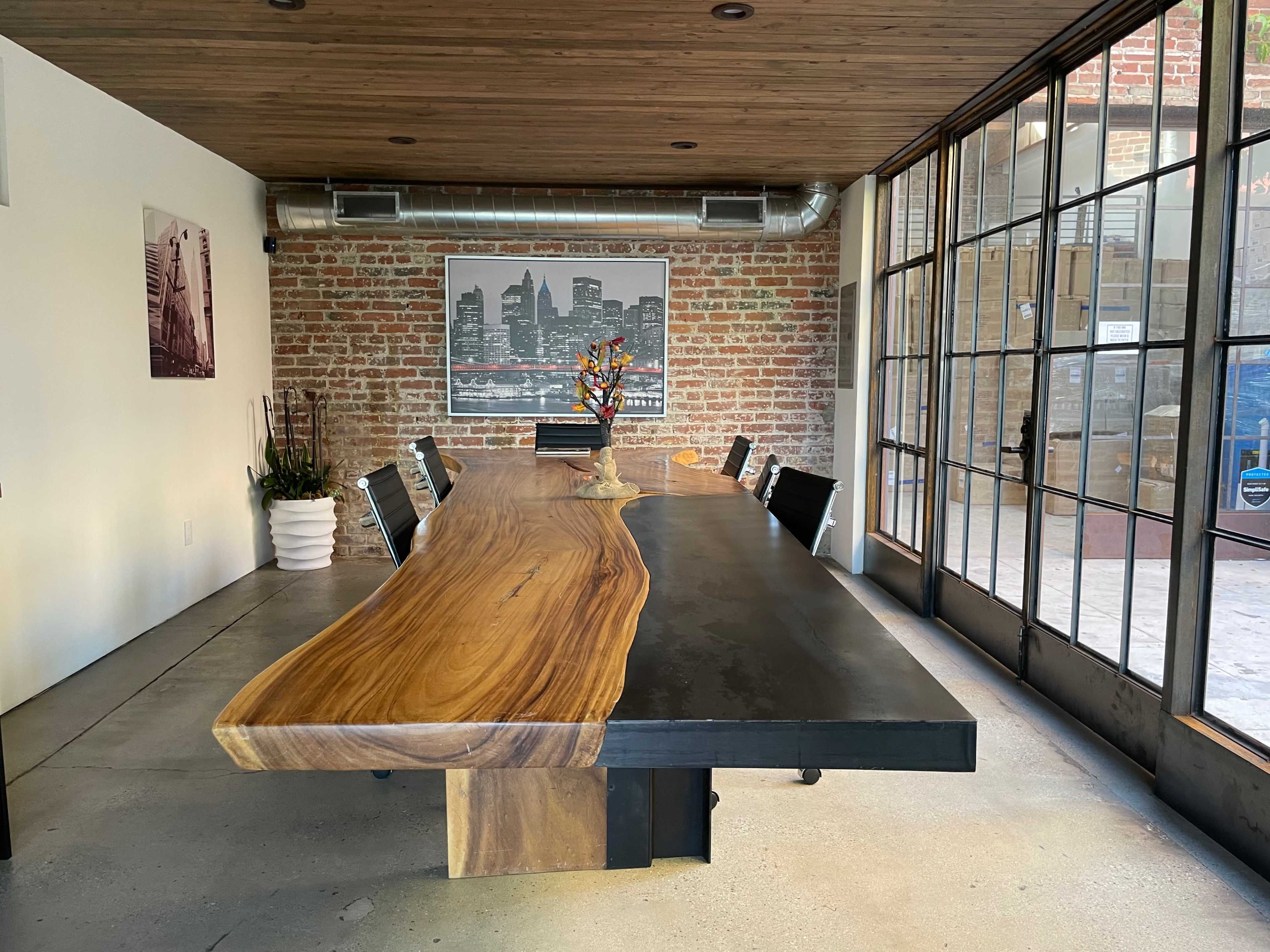 The image shows a modern conference room featuring a long wooden table with a black slab edge and black chairs, surrounded by brick walls and large glass doors.