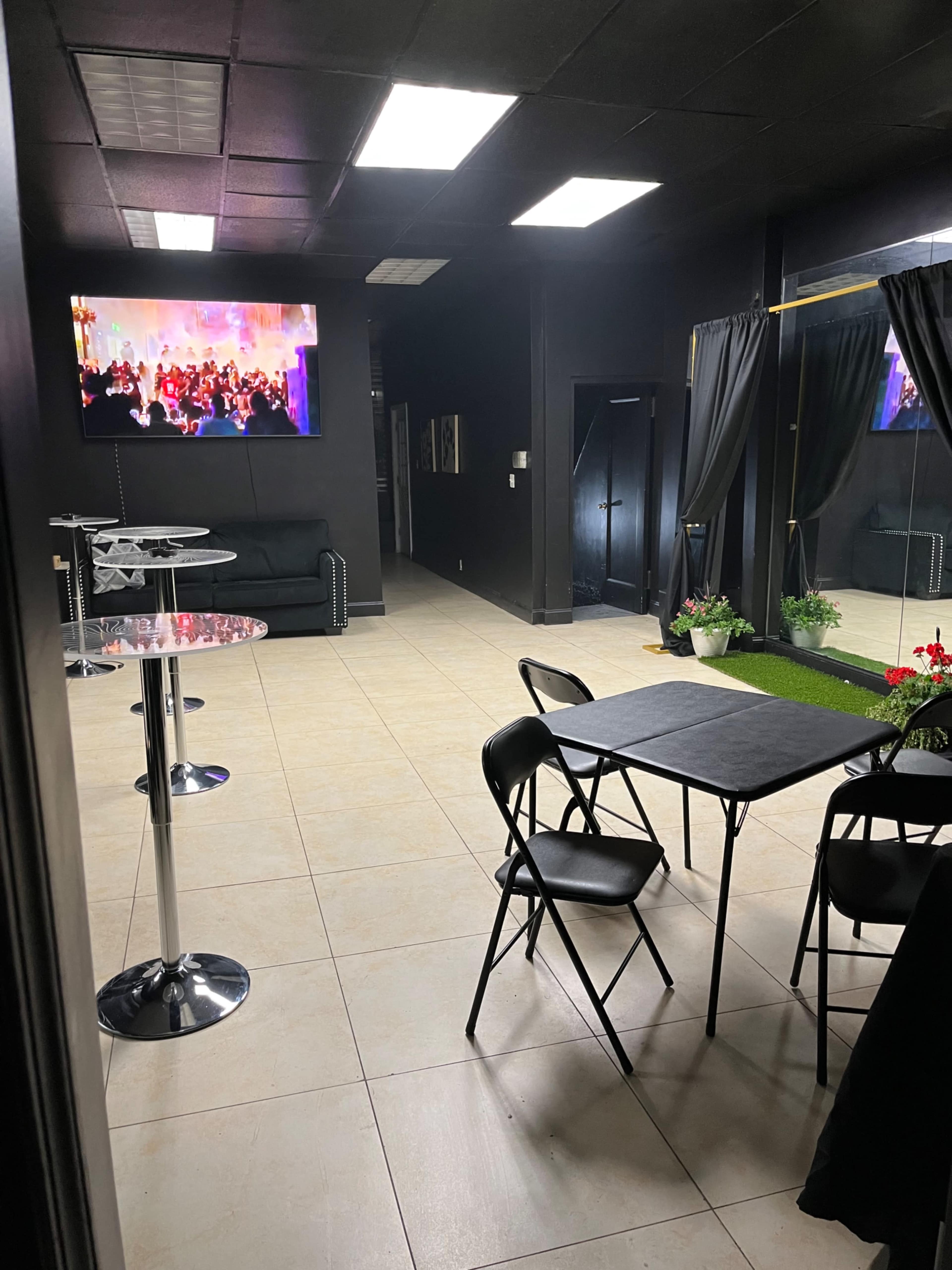 A modern interior space with a black ceiling and walls, featuring two tall tables with chairs, a large screen displaying a colorful event, and potted plants near a doorway.