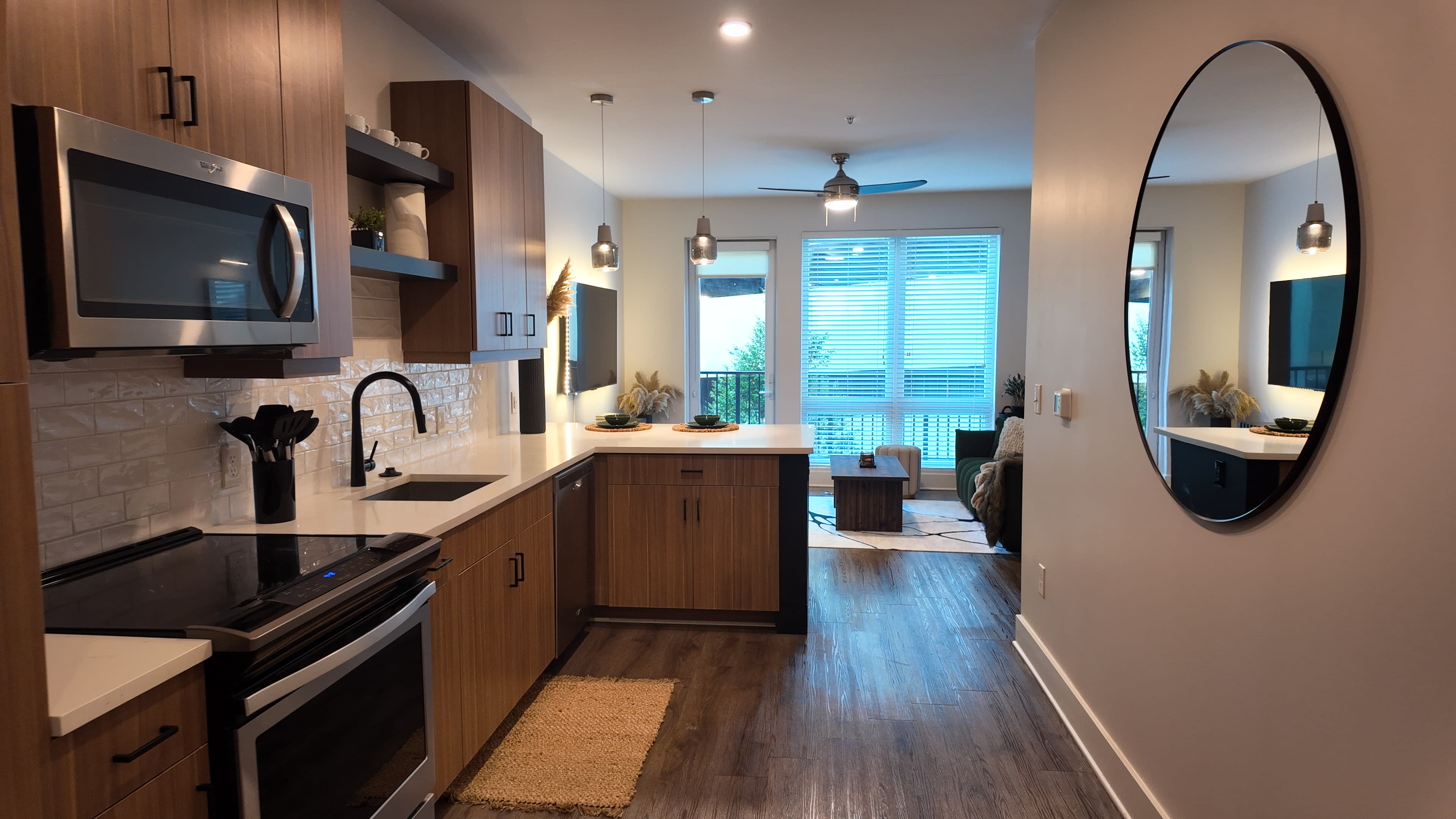 The image shows a modern kitchen area with wooden cabinets, stainless steel appliances, and a view into a living space that features a round mirror and natural light from a window.