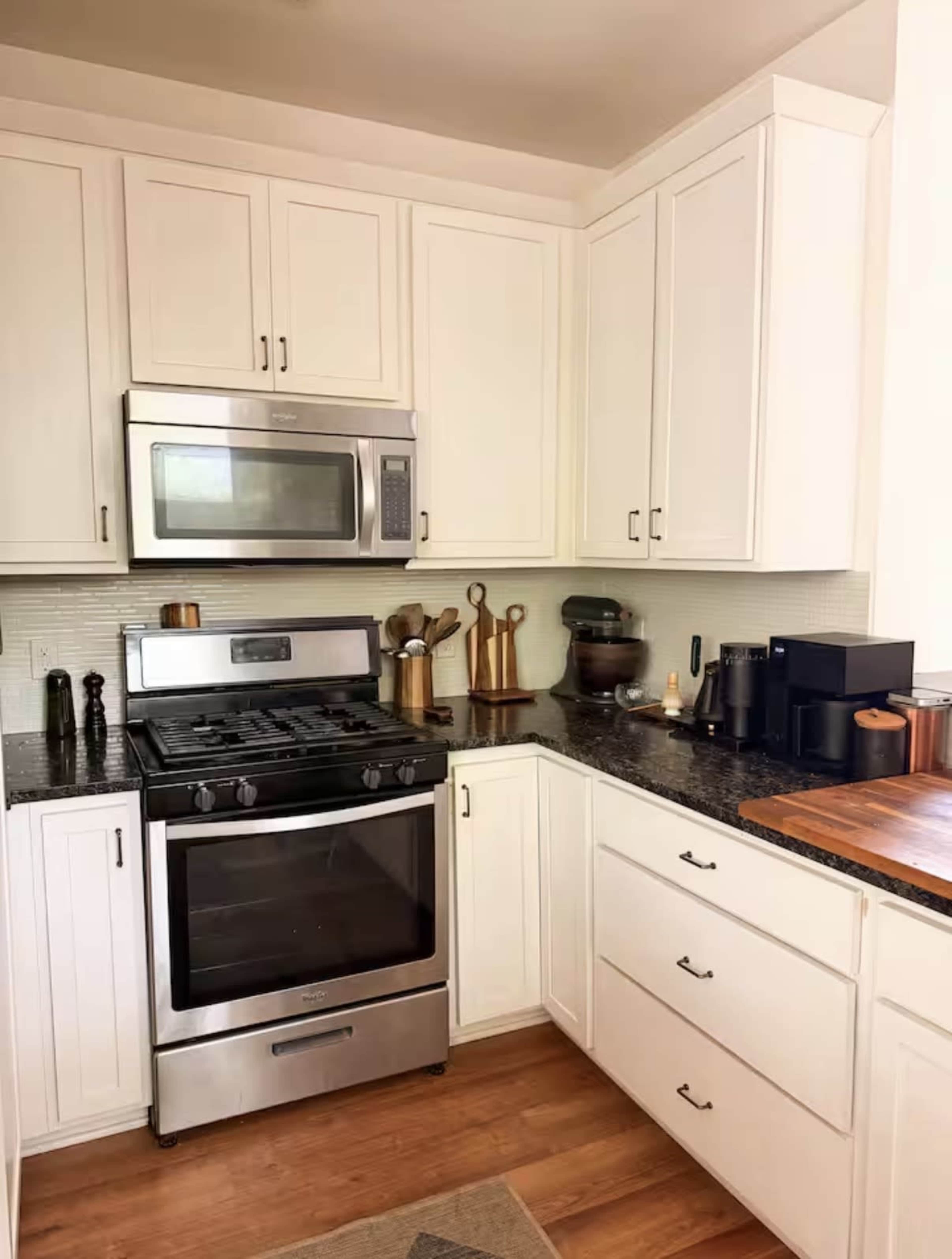 The image shows a compact kitchen with white cabinets, a stainless steel gas stove, a microwave above it, and a dark countertop featuring kitchen utensils and a coffee maker.