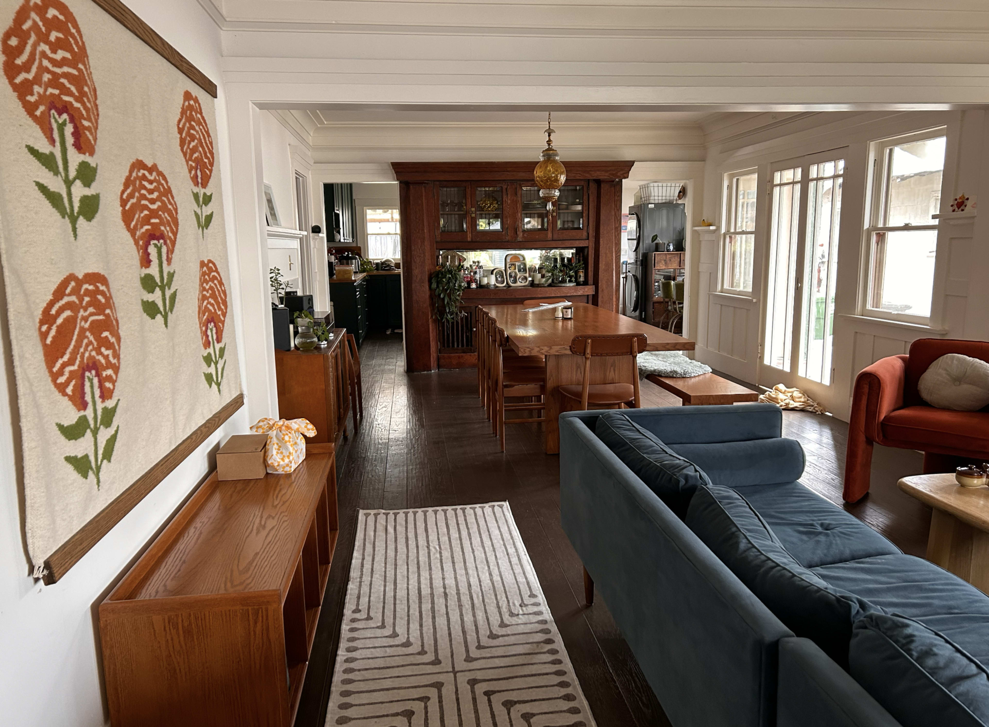 The image shows a spacious living area featuring a dining table, a wooden sideboard, and a mix of seating arrangements including a blue couch and an orange armchair.