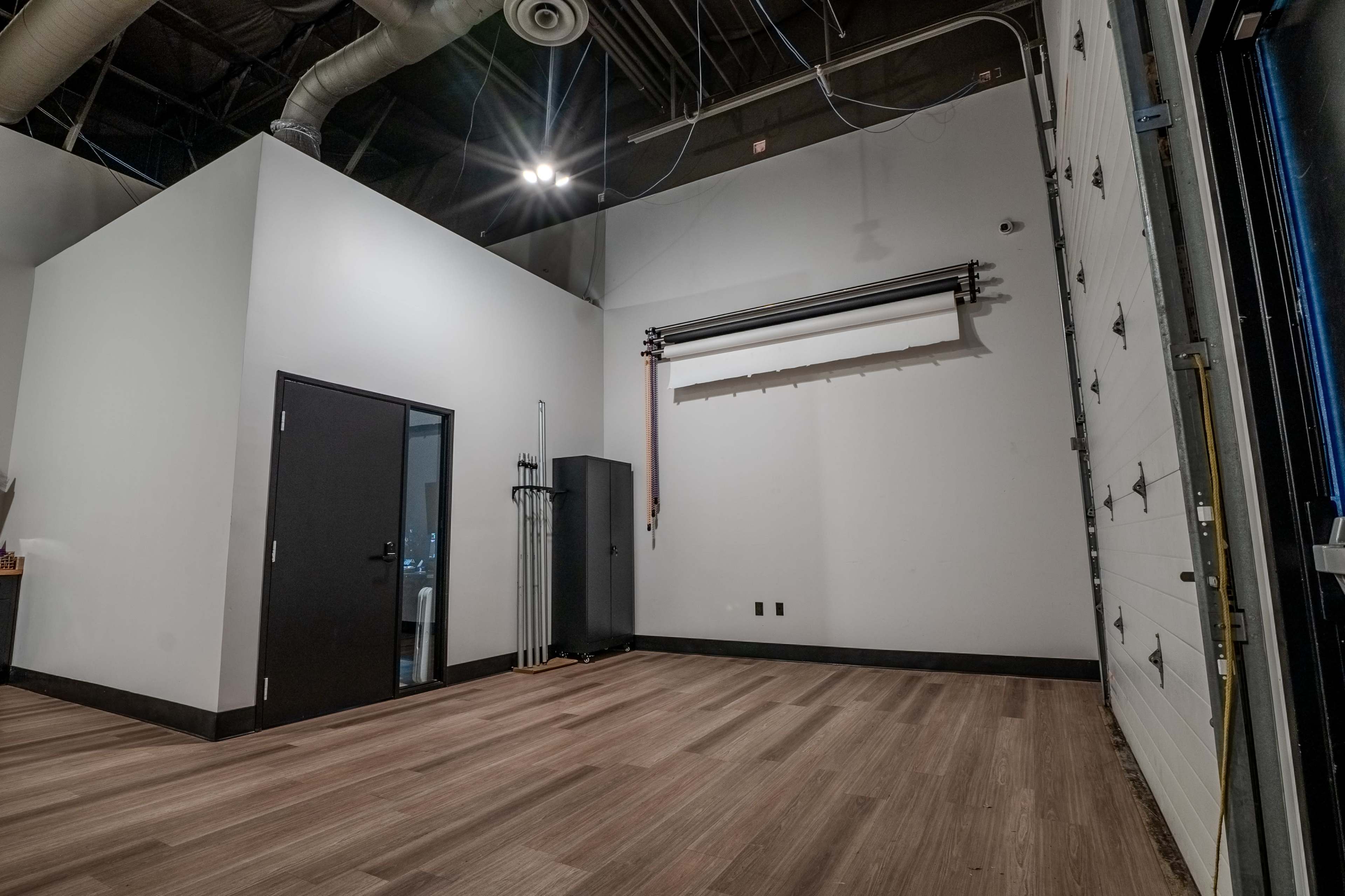 The image shows a spacious interior room with a door on the left, a roll-up door on the right, and wooden flooring.