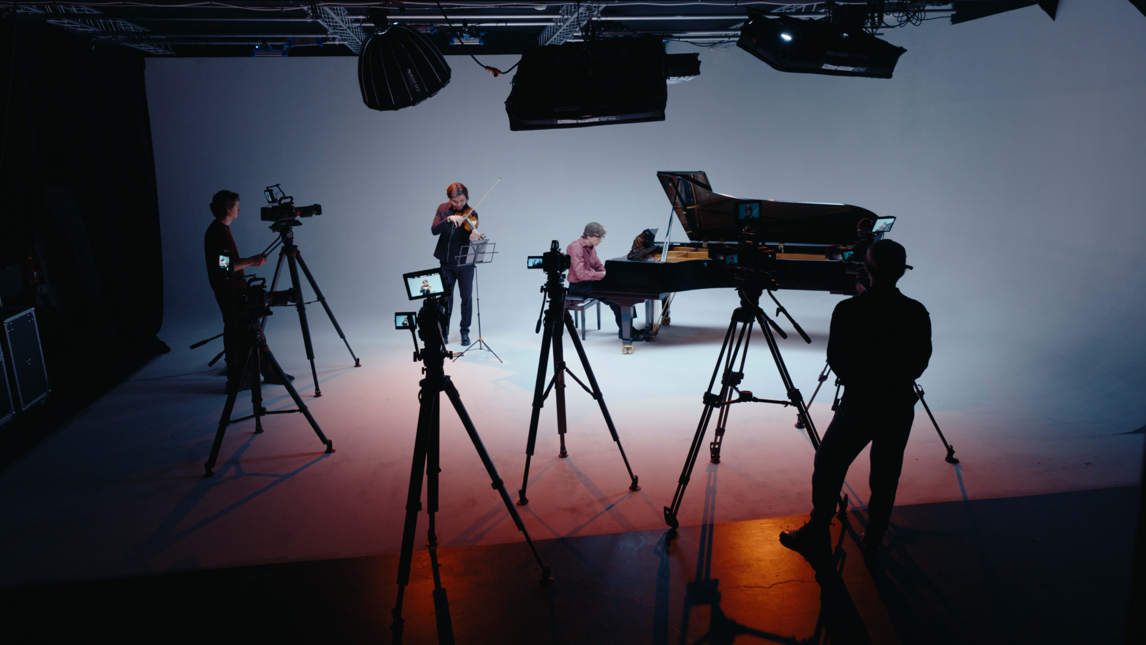 A pianist sits at a grand piano while a violinist performs next to them, and several cameras are set up around them in a studio setting.