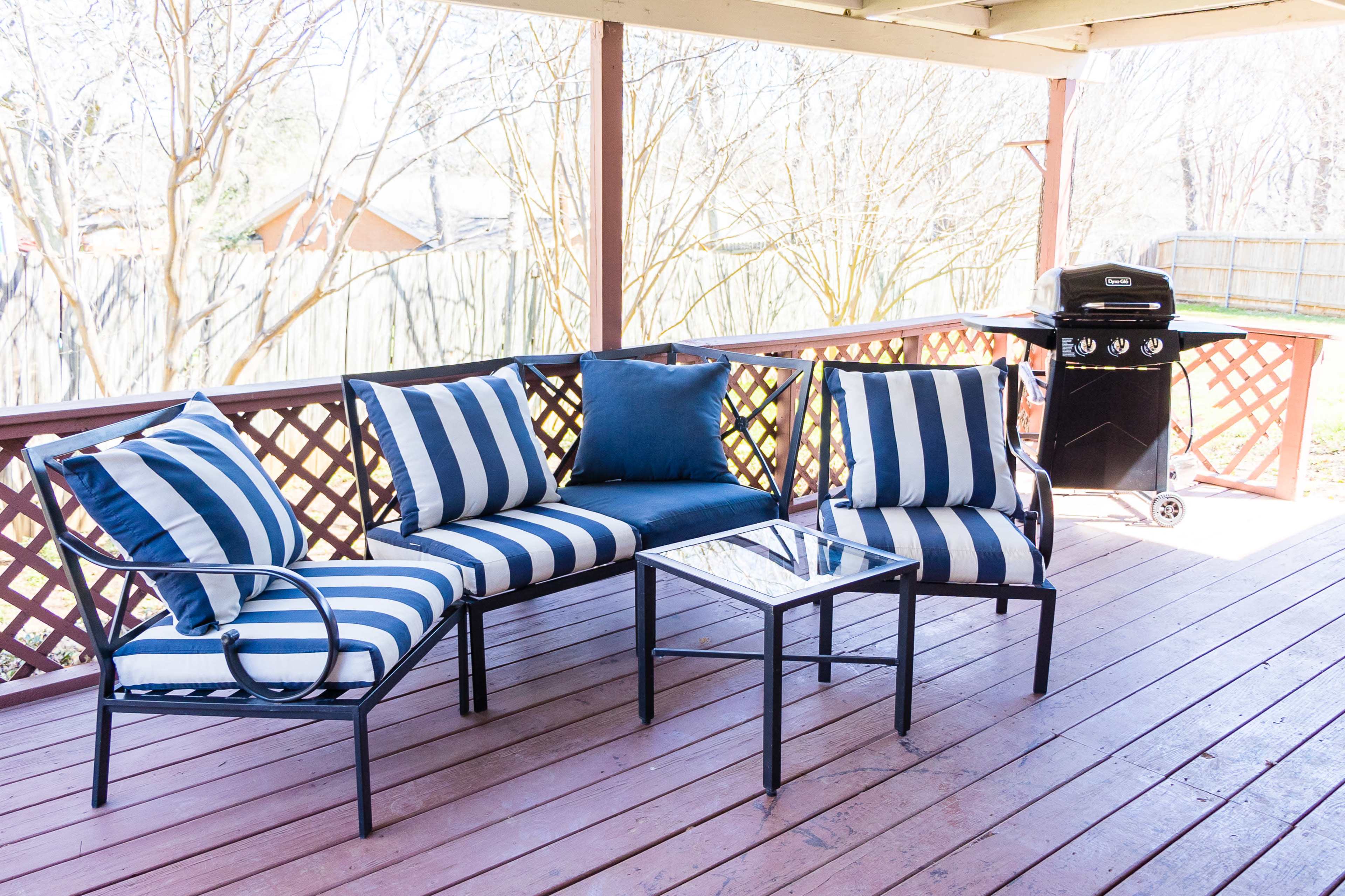 A patio area features a striped seating arrangement and a glass-top table next to a grill under a wooden pergola.
