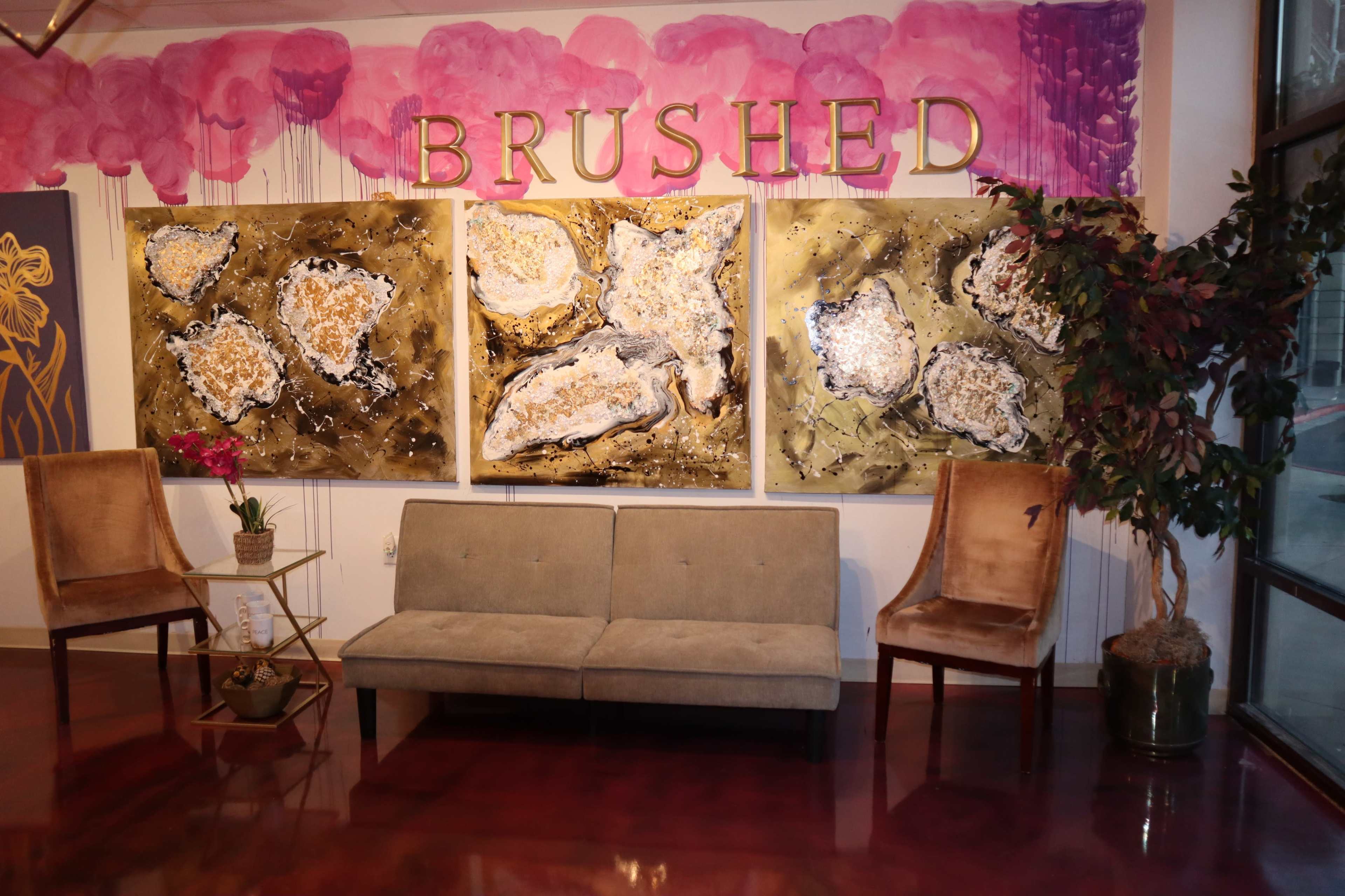 A lounge area with two chairs and a sofa positioned in front of three large abstract paintings, all set against a backdrop of pink decorative elements and the word "BRUSHED" in gold letters.