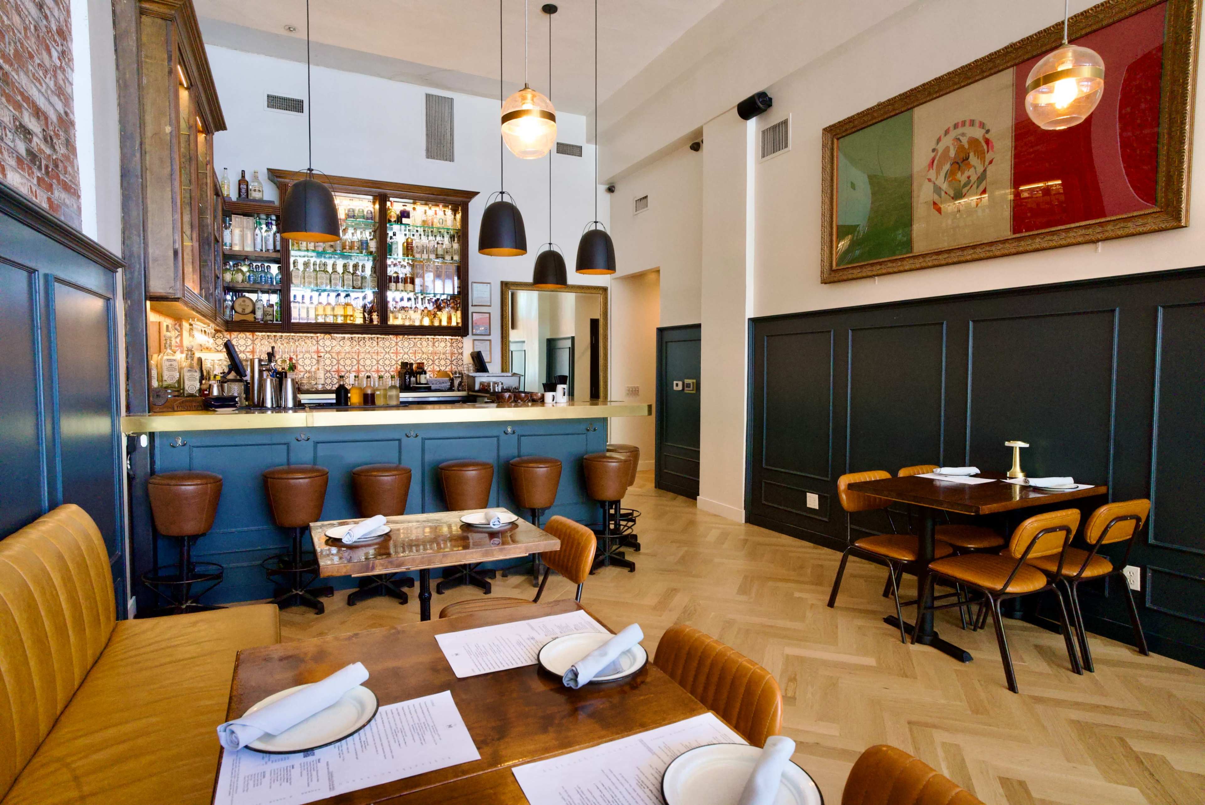 The image shows a stylish restaurant interior featuring a bar counter, leather seating, wooden tables, and decorative wall art.