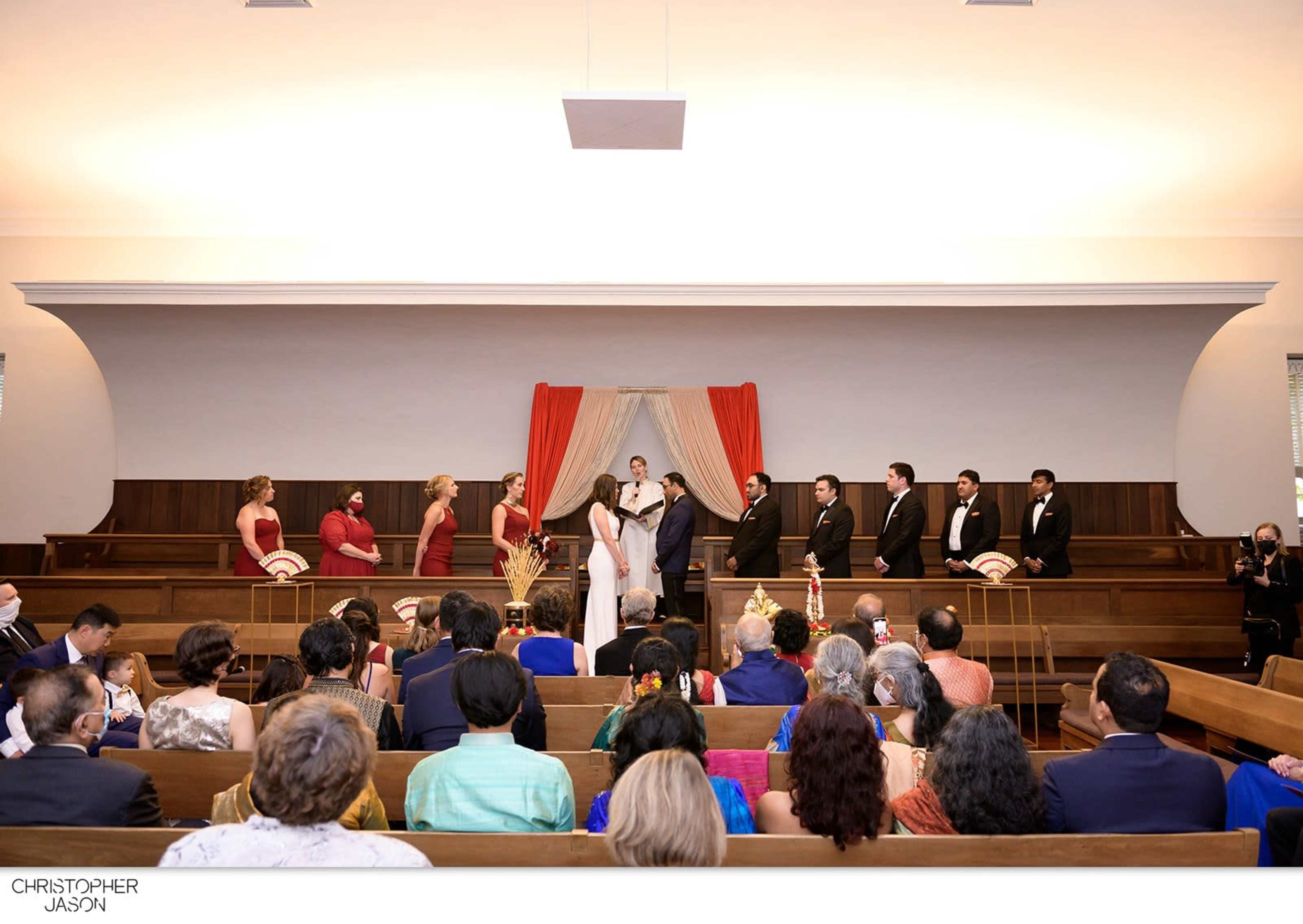 A wedding ceremony is taking place in a venue with a decorated altar, featuring a couple in the center and guests seated in wooden pews.