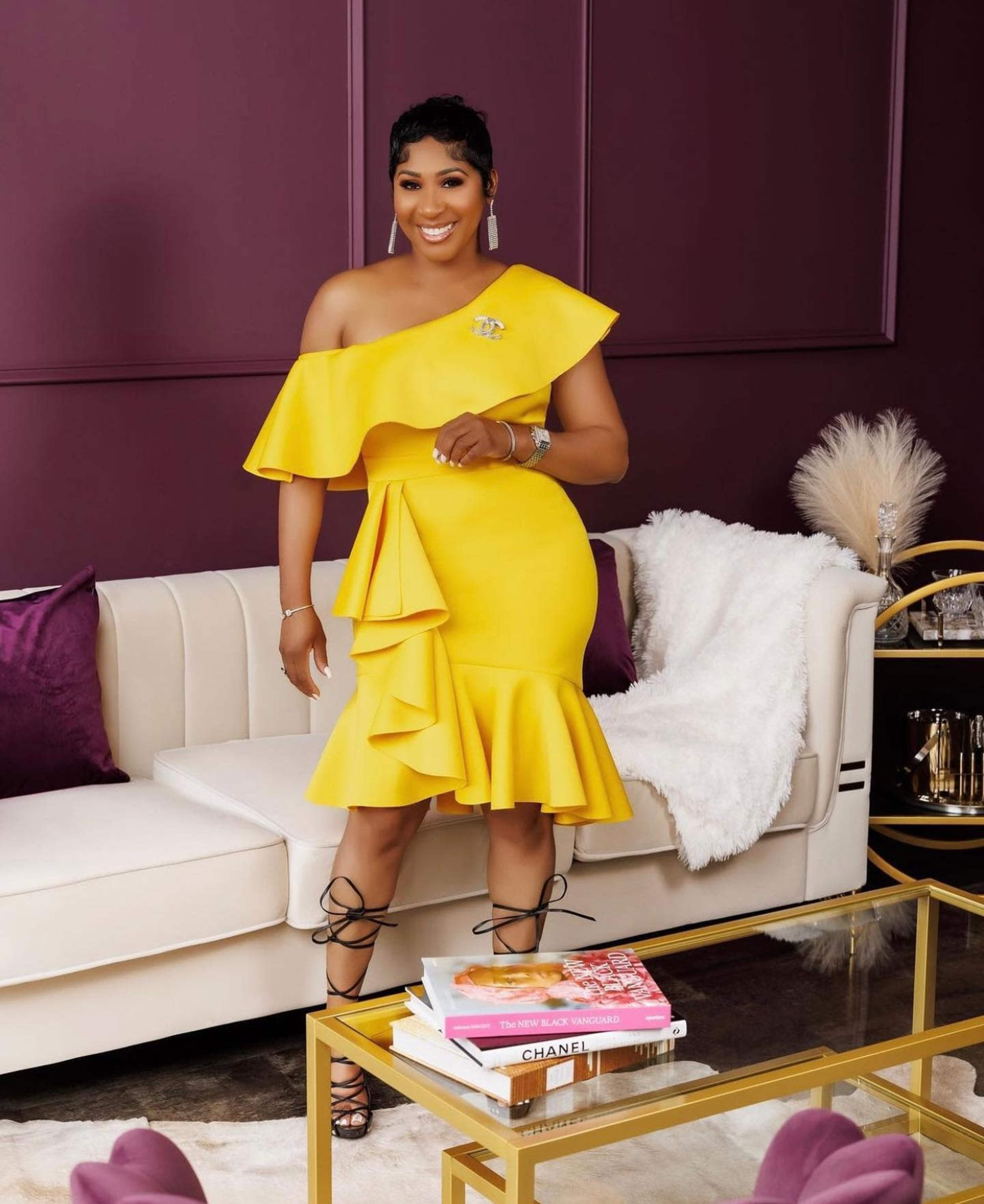 A woman in a bright yellow off-the-shoulder dress stands next to a beige sofa in a elegantly decorated room with dark purple walls and decorative cushions.