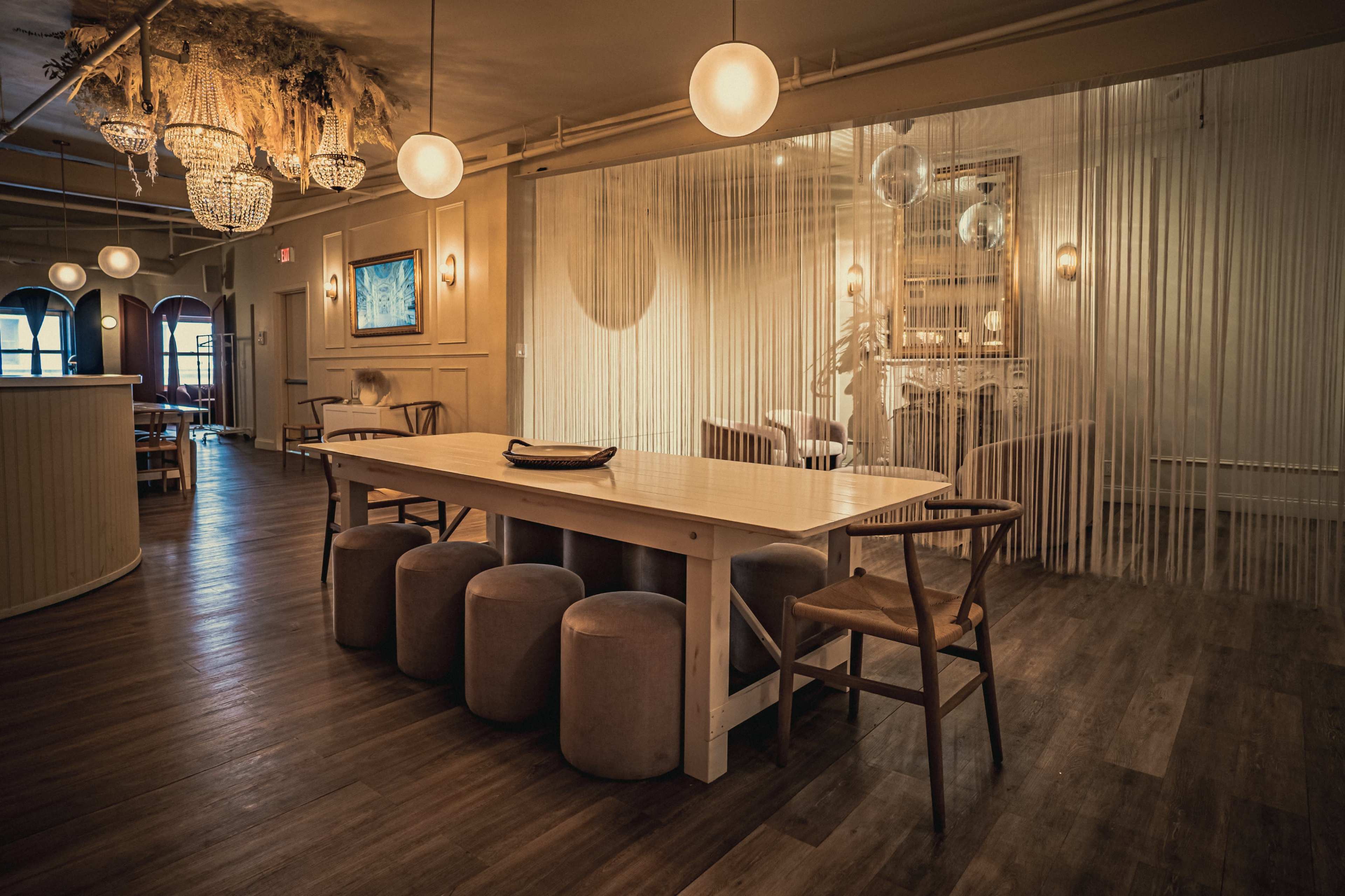 The image shows a modern dining area with a long wooden table surrounded by several stools and chairs, featuring warm lighting and decorative elements like hanging fixtures and a curtain.