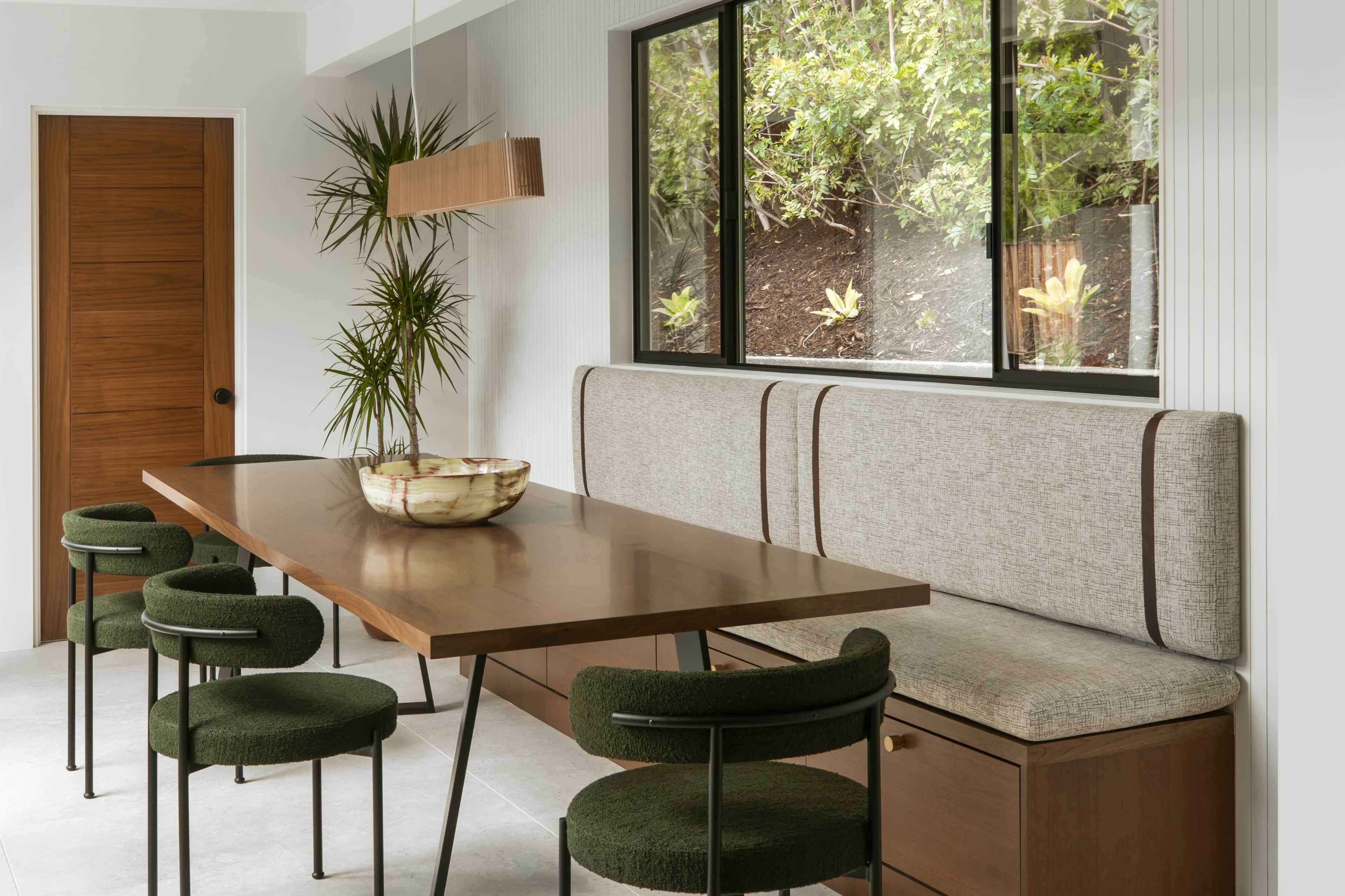 A modern dining area features a long wooden table with a built-in bench and green chairs near large windows overlooking greenery.