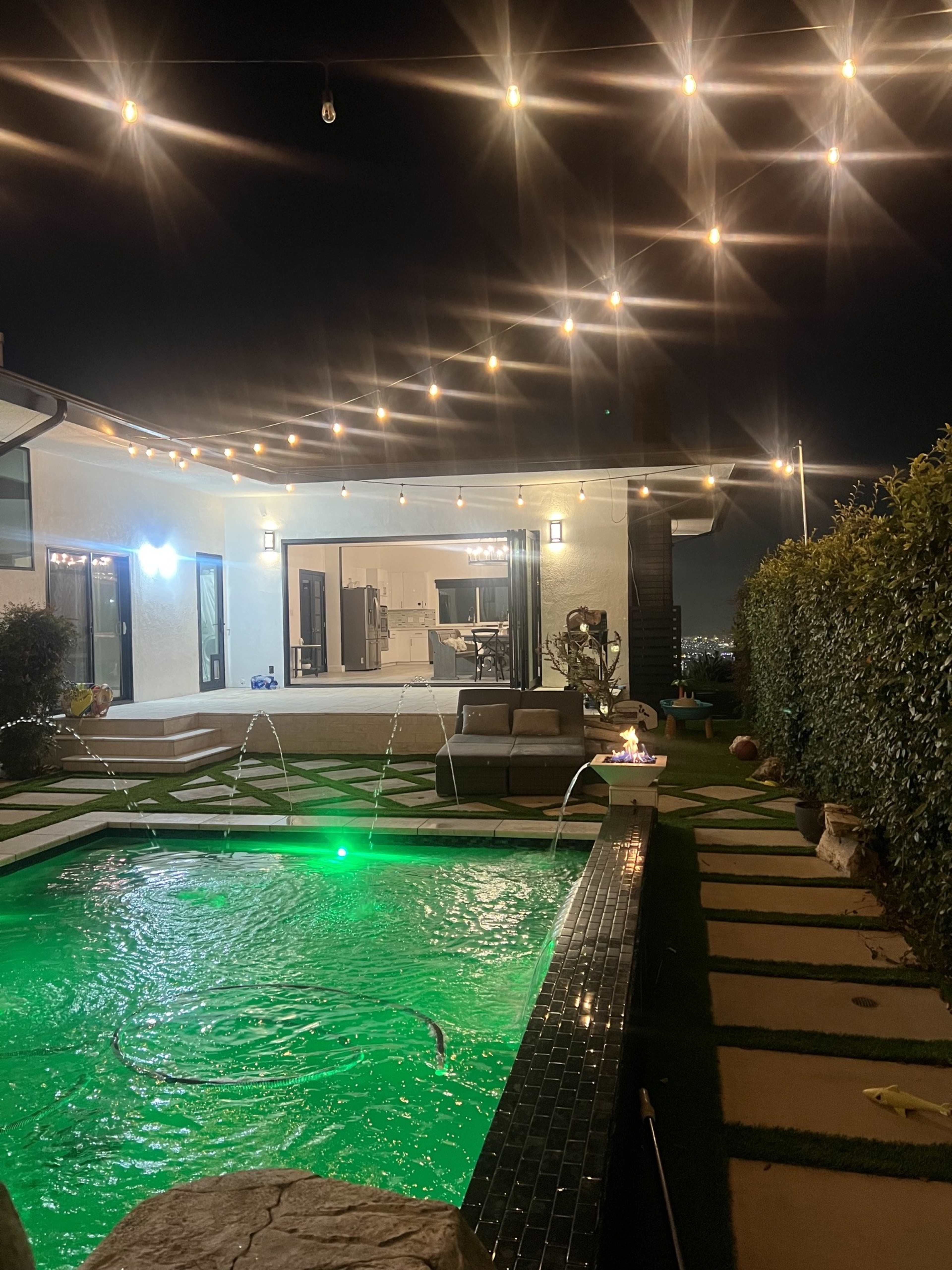 A brightly lit backyard features a pool with green illumination, surrounded by illuminated string lights and a modern house with large windows in the background.