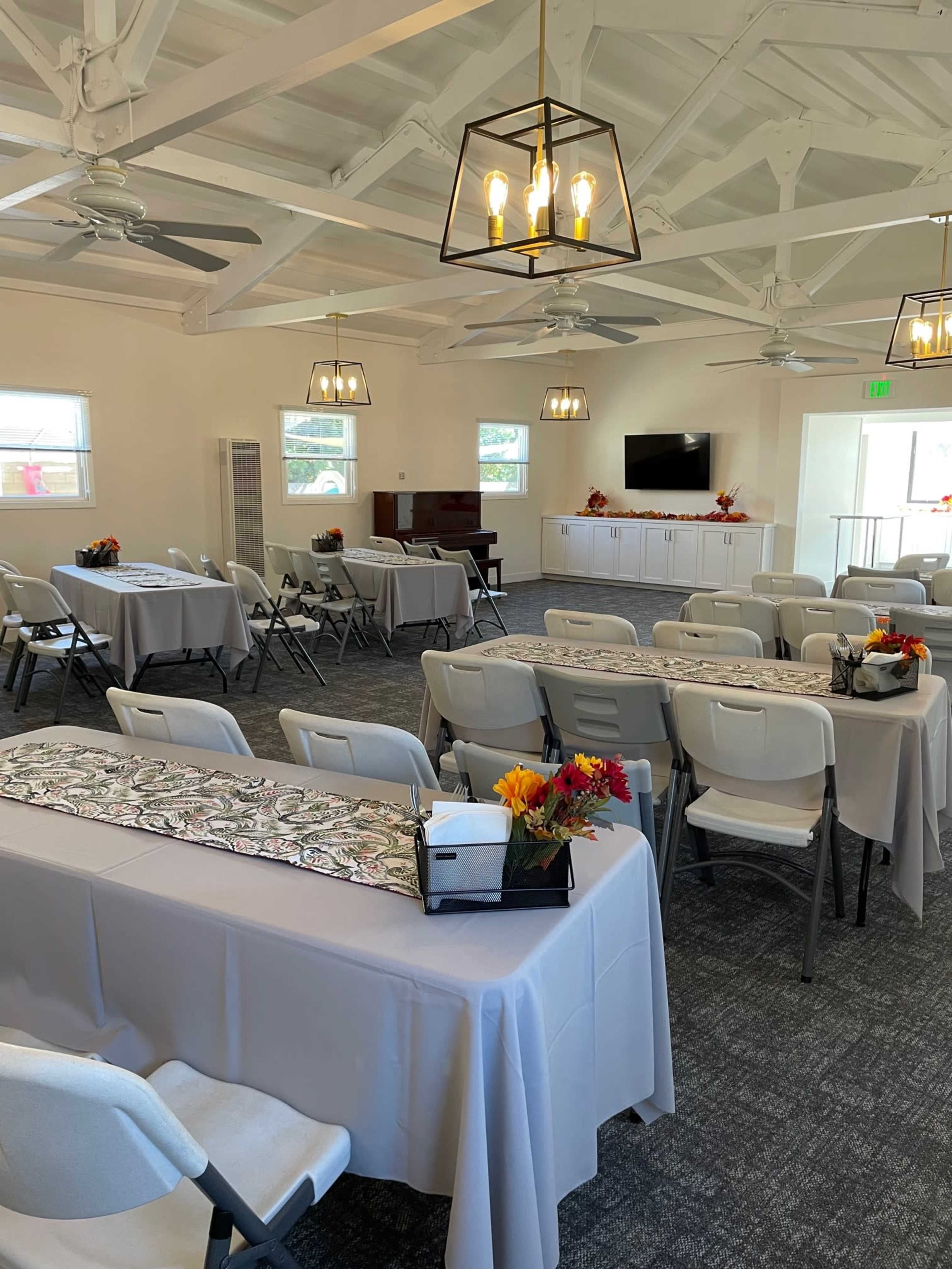 A spacious event room features several tables set with white tablecloths, chairs, and decorative centerpieces, under bright lighting and a high ceiling with exposed beams.