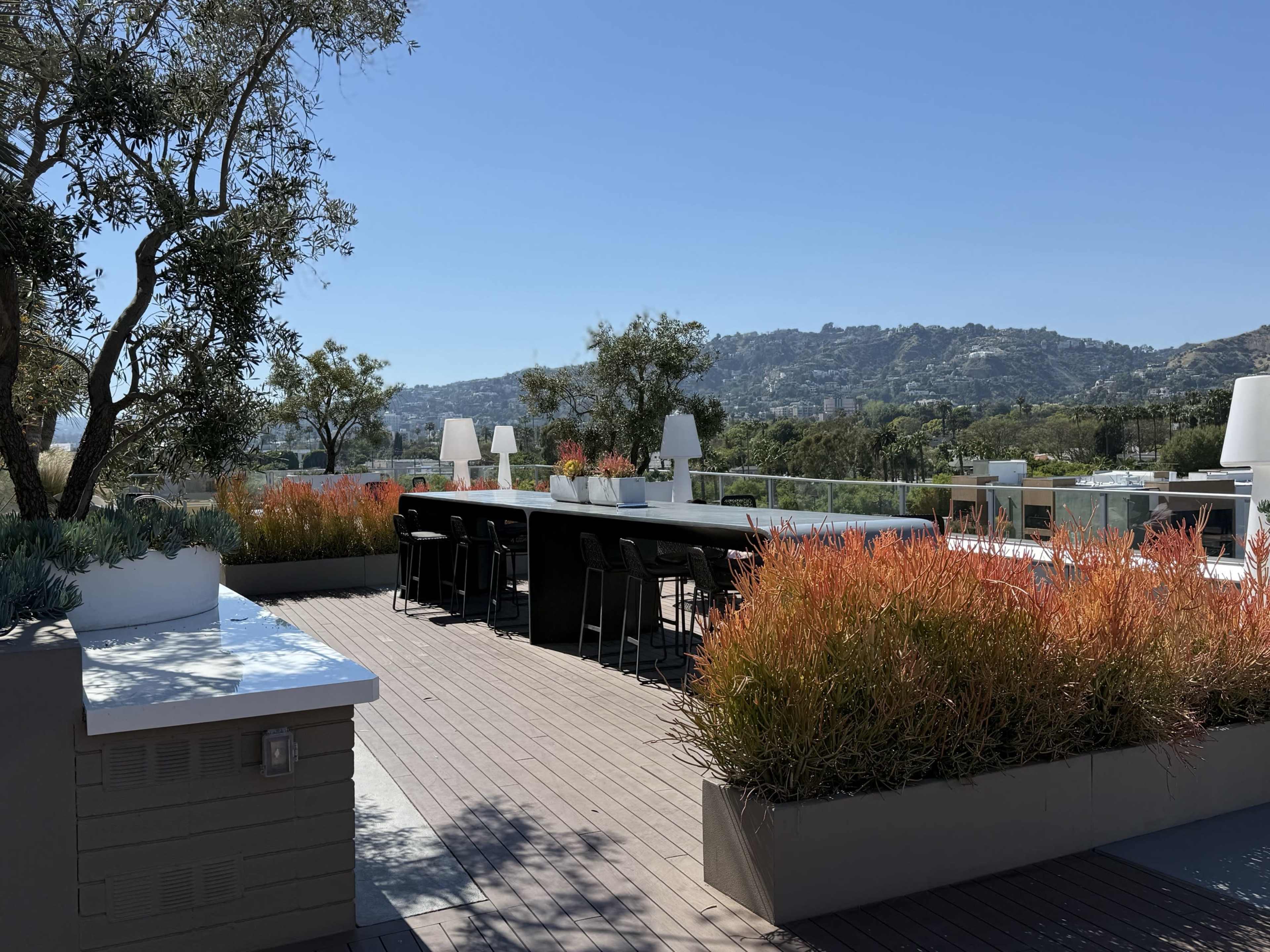A rooftop terrace features a long table surrounded by chairs, with greenery and colorful plants along the edges, set against a hillside view under a clear blue sky.