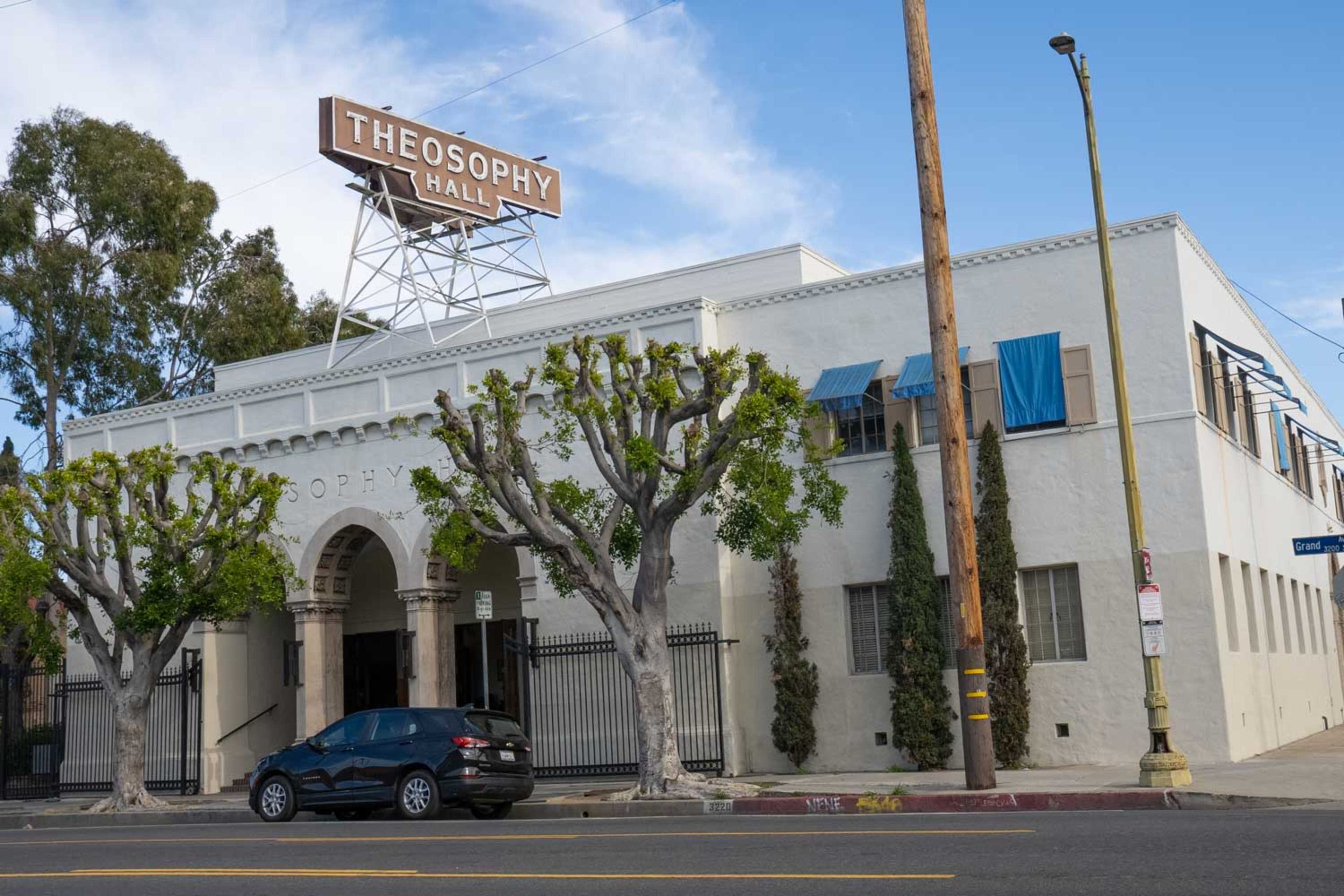 The image shows a white building with arched entrances labeled "Theosophy Hall," flanked by trimmed trees and blue window coverings, under a clear sky.